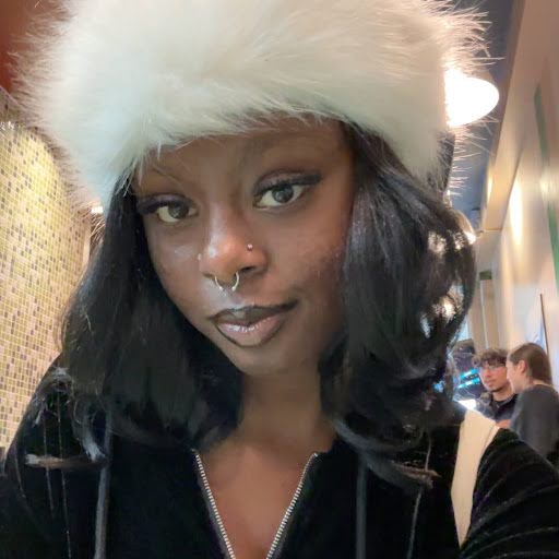 Naya is a dark-skinned woman with long dark brown hair. She is wearing a fur cap and a black velour hoodie.