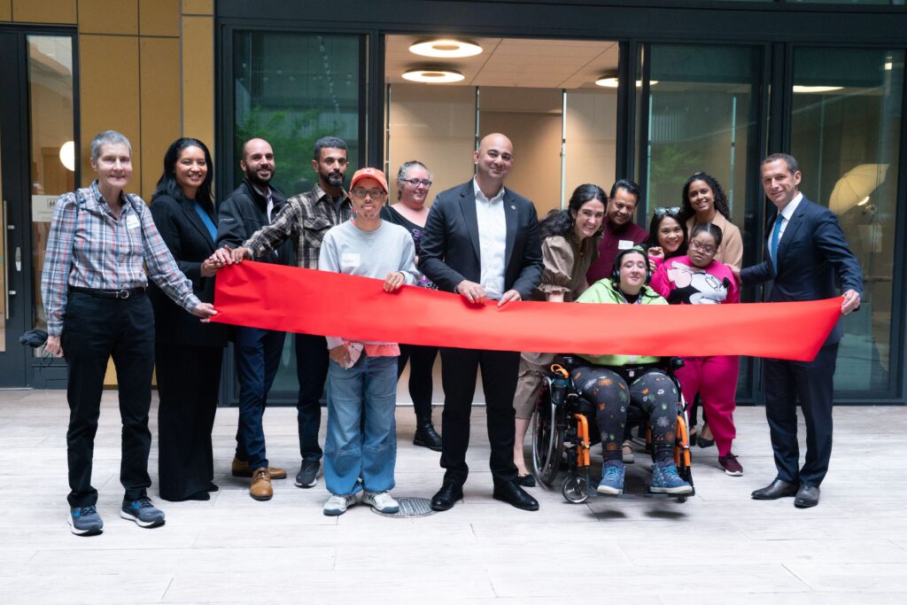 A diverse group of people, including people with and without disabilities, stand together smiling during a ribbon-cutting ceremony. Two people hold a long red ribbon across the front, and one person in a wheelchair is near the center of the group. The group is standing in an indoor courtyard area in front of large glass doors.