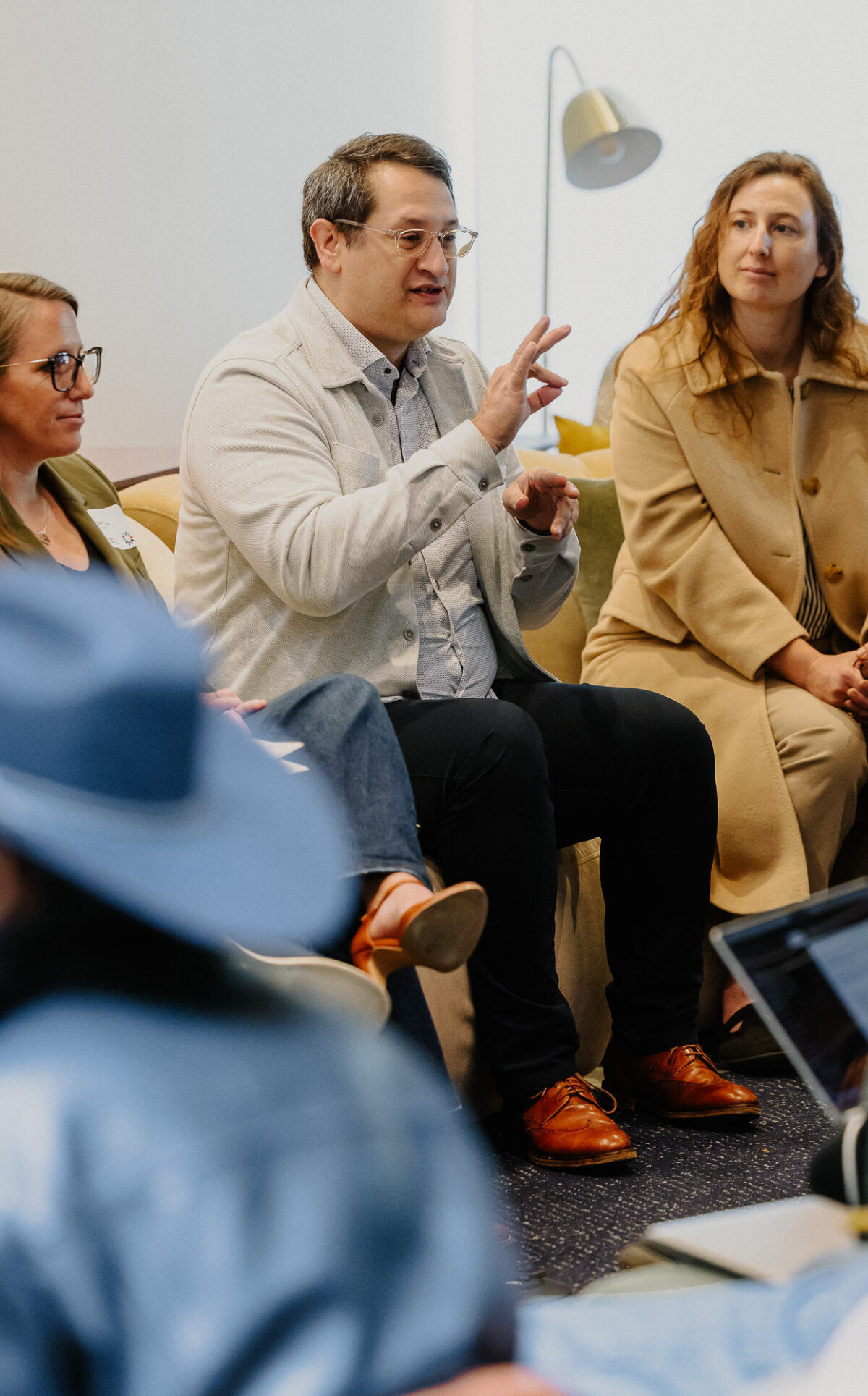 Anne Riggs (David Baker Architects), Sarah Vaccaro (Architects FORA), and Jeffrey Mansfield (MASS Design) are seen sitting on a couch as they speak on a panel. Erick Mikiten (Mikiten Architecture), a wheelchair rider, is seen sitting next to the couch,