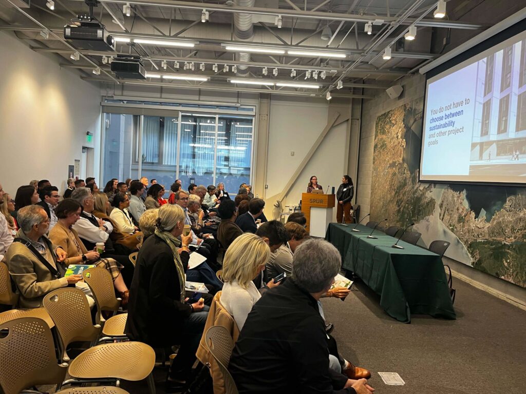 A large audience sits in rows inside a modern conference room as Louisa and Fiona stand at a podium at the front of the room. A slide on the screen reads, “You do not have to choose between sustainability and other project goals.”