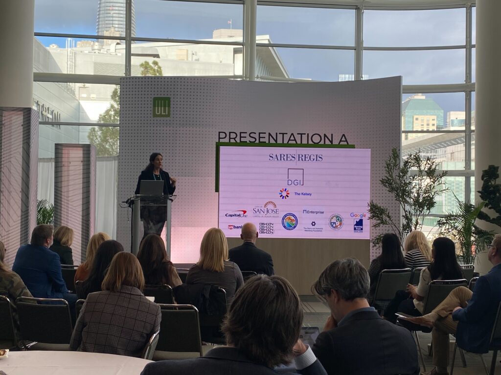 Micaela, a white woman with long brown hair, stands on a stage with a laptop and a microphone, speaking to an audience. Behind her is a large screen displaying partner logos, including The Kelsey, Sares Regis, DGI, and others. Large windows show downtown San Francisco in the background.