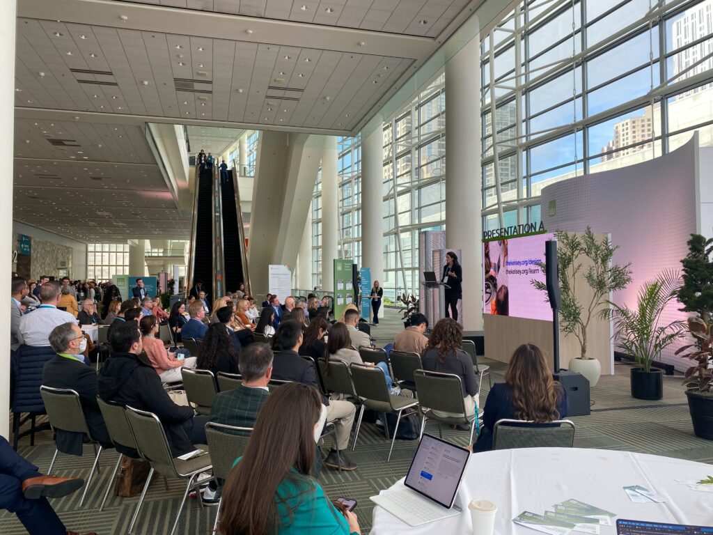 Micaela speaks on a stage in a spacious conference hall filled with attendees. A large screen behind her displays The Kelsey’s “Learn Center” and the “Inclusive Design Standards” website links. An escalator and tall windows are visible in the background.