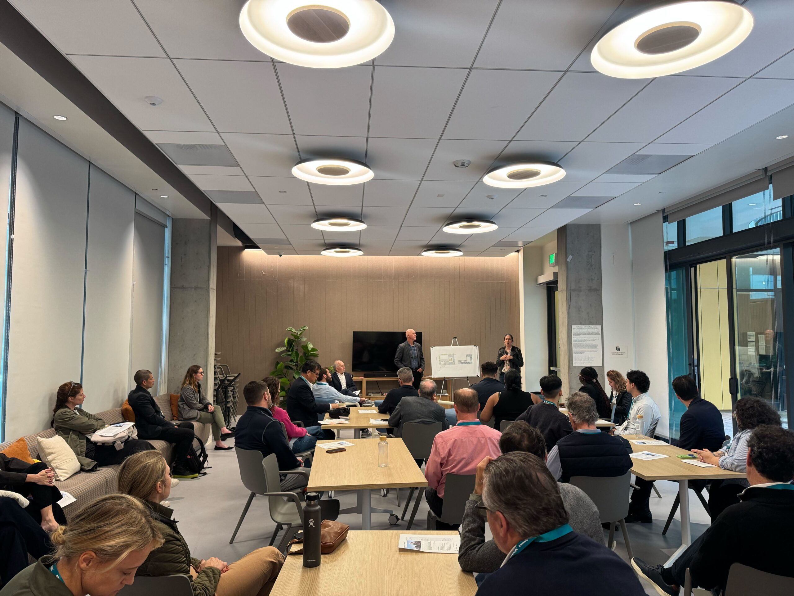 A group of attendees sits in The Kelsey Civic Center community room as Louisa stands at the front next to an easel with architectural drawings. People are seated around tables and along the walls, listening to the presentation.