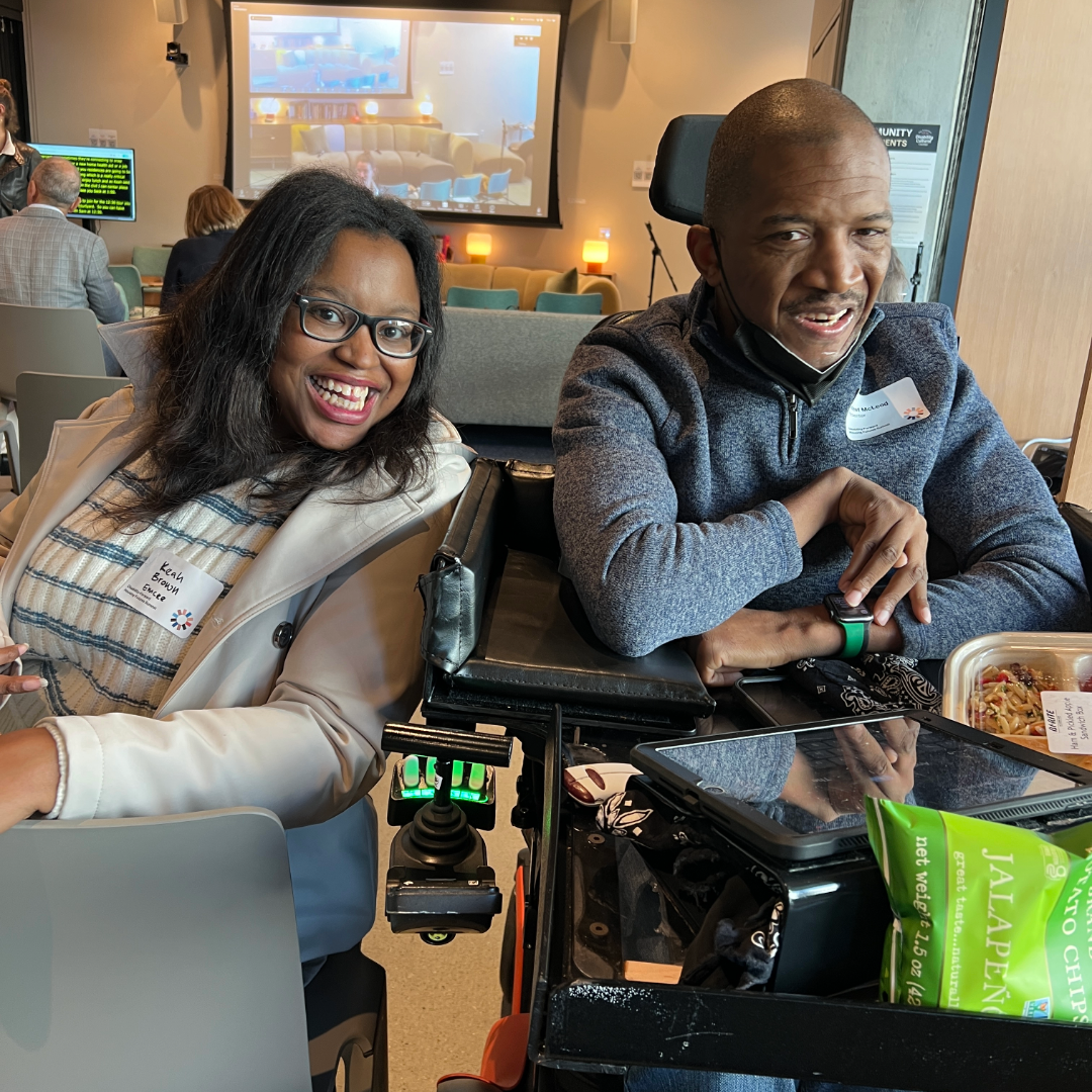 Two people smile while sitting side by side. One person is using a power wheelchair with a tray in front, and the other is sitting next to them wearing glasses and a light-colored coat. A presentation screen is visible in the background with people seated in a cozy, well-lit room.