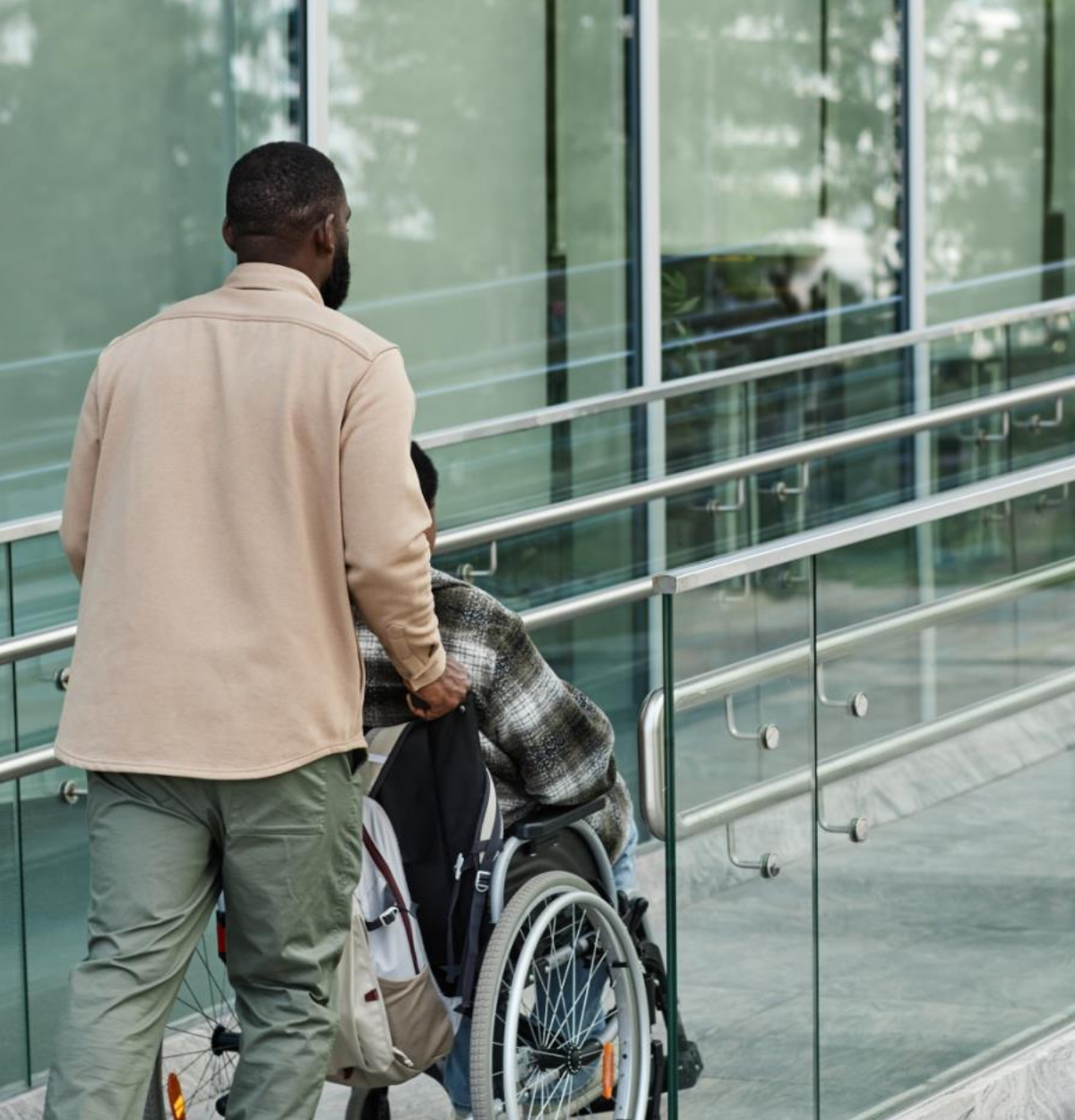 A person pushes another person using a wheelchair up a ramp outside of a modern glass building.
