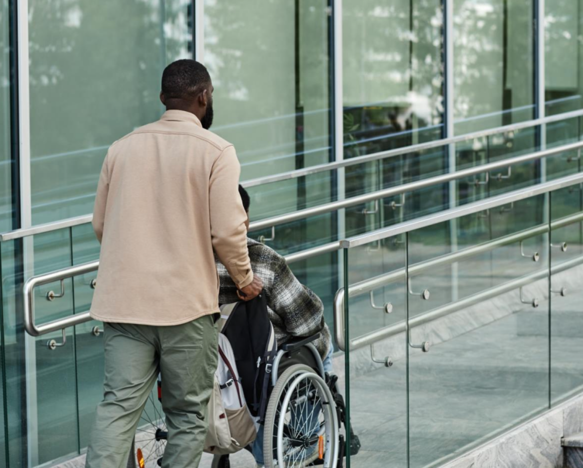 A person pushes another person using a wheelchair up a ramp outside of a modern glass building.