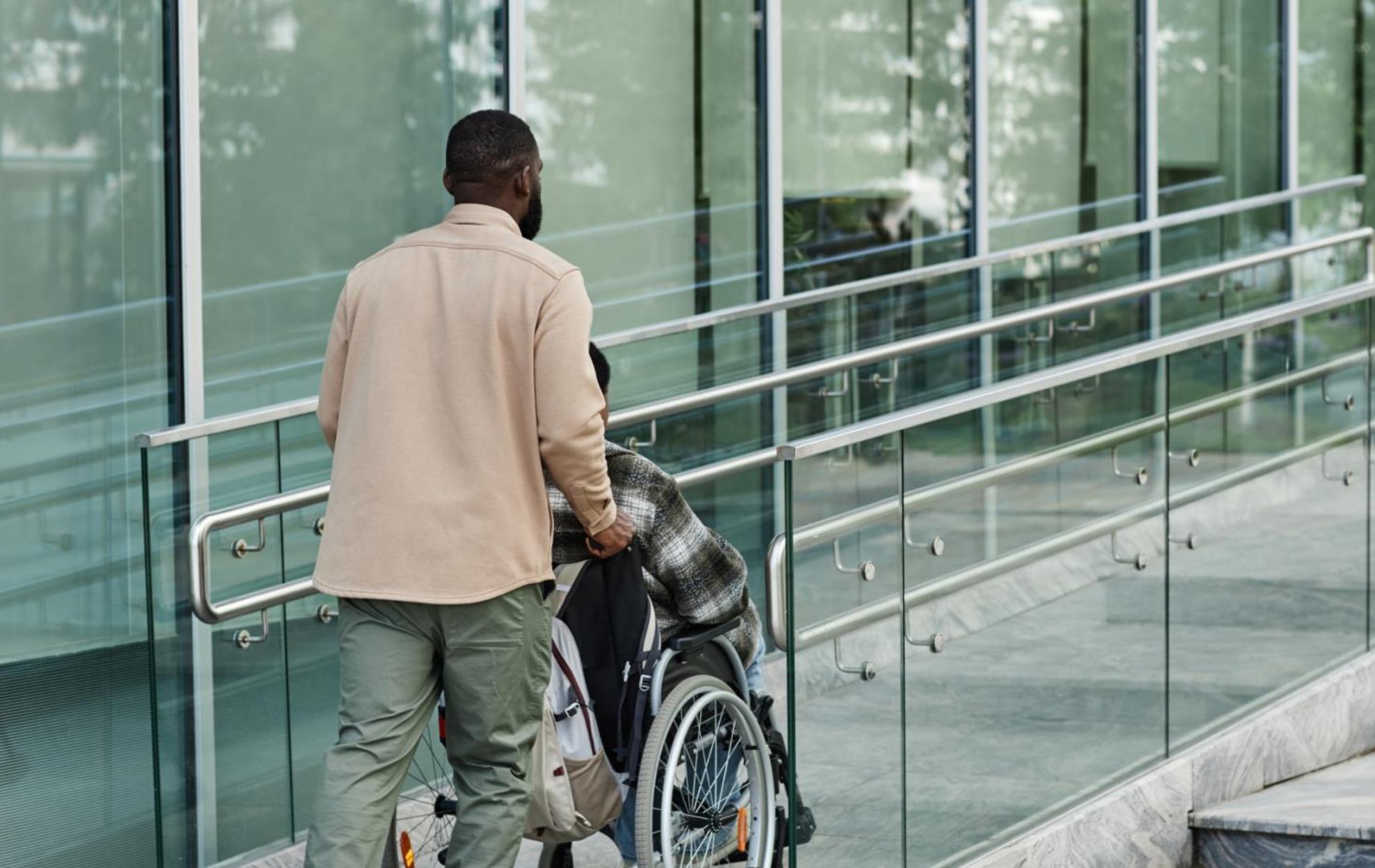 A person pushes another person using a wheelchair up a ramp outside of a modern glass building.