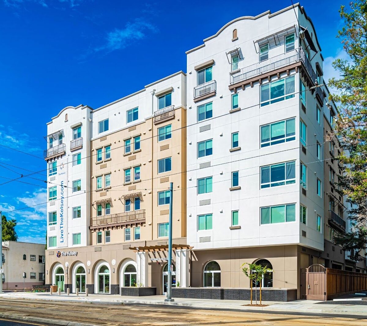 The exterior of The Kelsey Ayer Station, a light beige and white apartment building with large windows, small balconies, and arched ground-floor entrances. A banner reading ‘LiveTheKelsey.com’ hangs along one side. The building sits along a street with rail tracks, trees, and a bright blue sky overhead.”