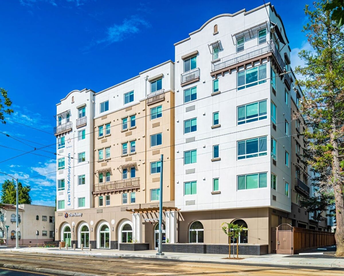 The exterior of The Kelsey Ayer Station, a light beige and white apartment building with large windows, small balconies, and arched ground-floor entrances. A banner reading ‘LiveTheKelsey.com’ hangs along one side. The building sits along a street with rail tracks, trees, and a bright blue sky overhead.”