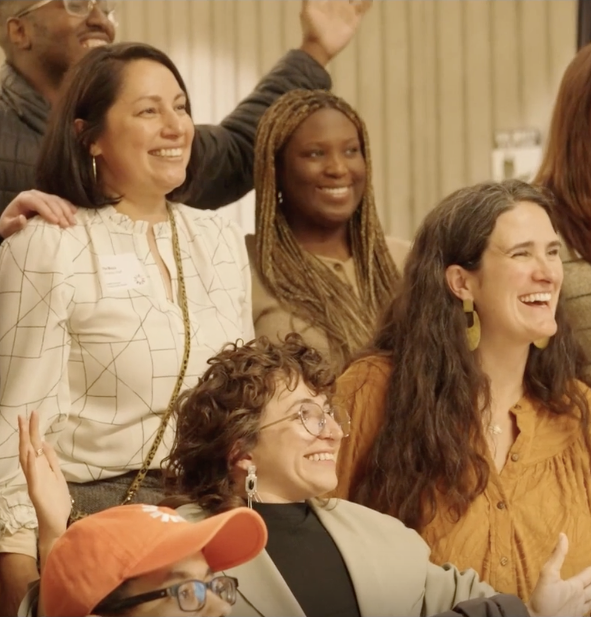 A diverse group of adults with and without disabilities, smiling and laughing as they pose for a group photo. They are relaxed and celebratory, with some people raising their hands or placing their arms around each other.