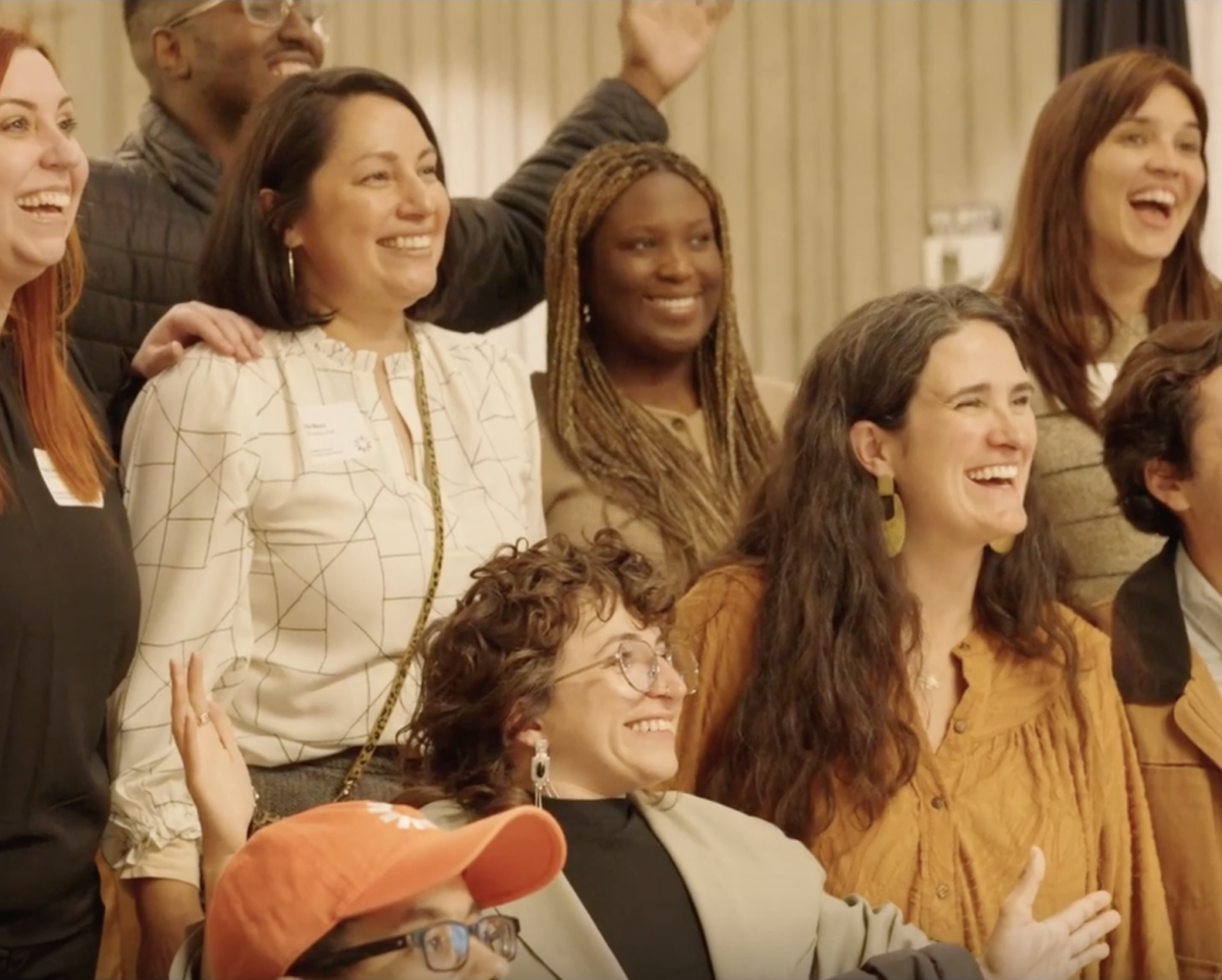 A diverse group of adults with and without disabilities, smiling and laughing as they pose for a group photo. They are relaxed and celebratory, with some people raising their hands or placing their arms around each other.