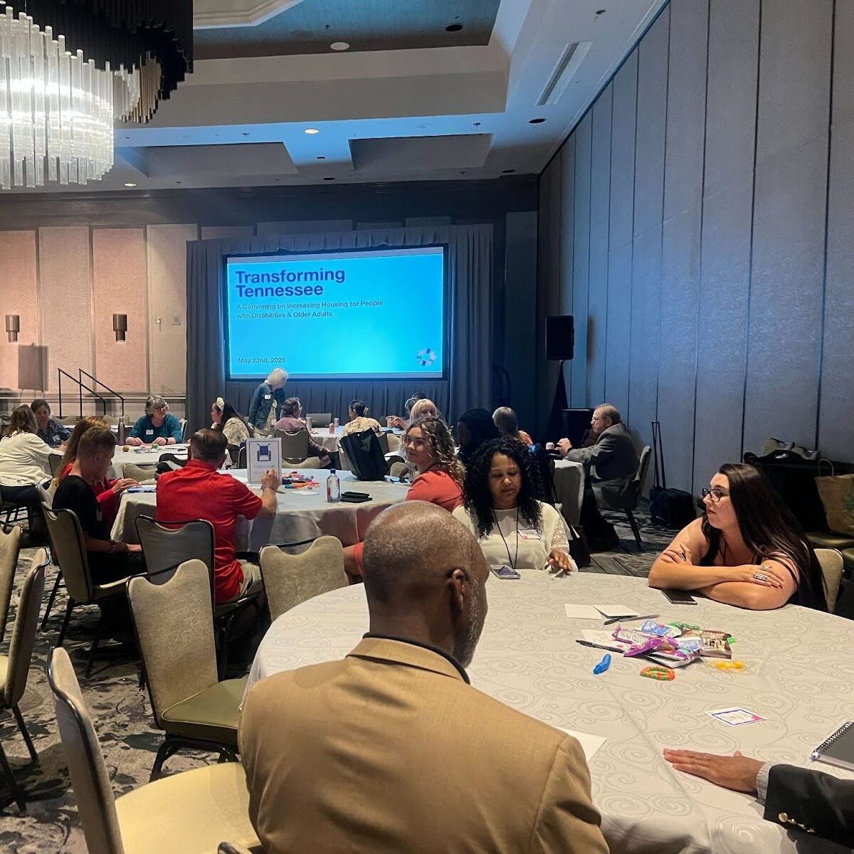 People sit in a conference room at round tables. On a screen in front of the room, a presentation with the words, “Transforming Tennessee,” is visible.