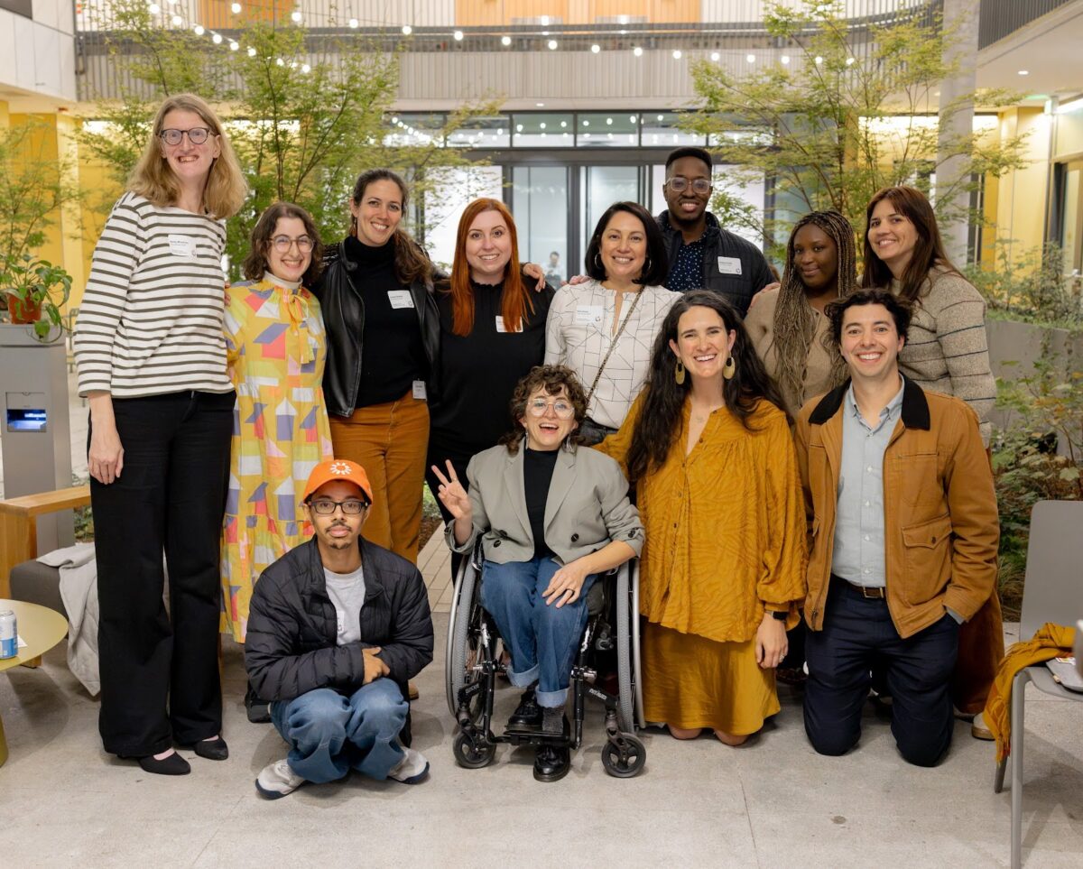 A diverse group of adults with and without disabilities, smiling and posing for a group photo. They are in the courtyard of The Kelsey Civic Center: string lights, greenery and seating is visible around them.