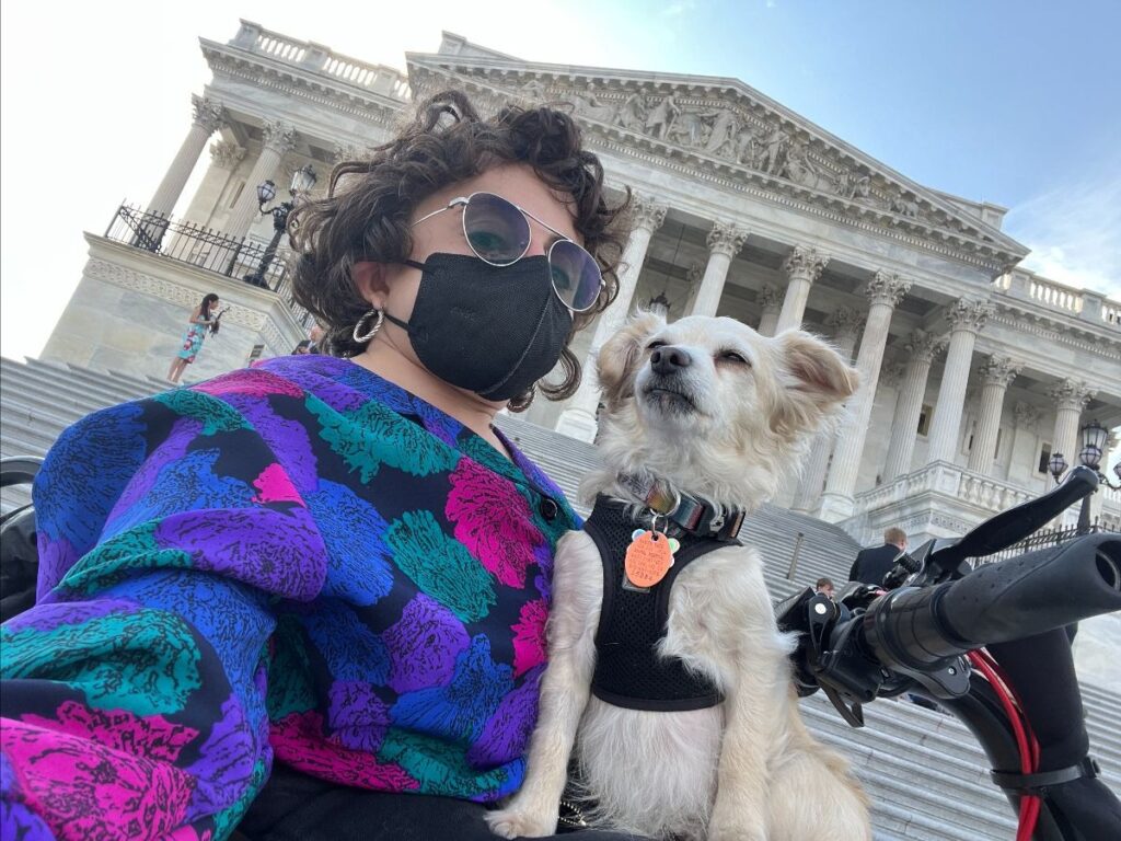 Allie, our Managing Director, takes a selfie in front of Congress. They are a white queer person in a scooter, wearing sunglasses and holding a dog on their lap. They are wearing a black mask and a purple, blue, and pink sweater. 