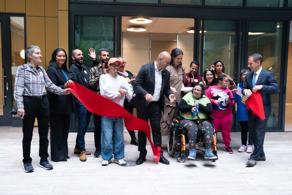 Image description: A diverse group of people gathers for a ribbon-cutting ceremony in front of The Kelsey Civic Center’s courtyard entrance. A person in a wheelchair holds scissors after cutting a bright red ribbon while others cheer and smile around them. The group includes people of different ages with and without disabilities, wearing casual and professional attire.