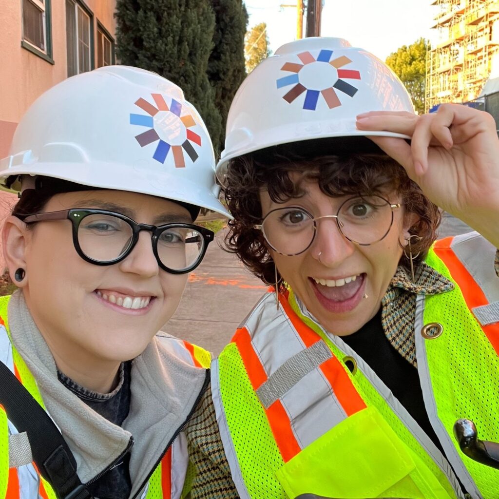 A picture of The Kelsey’s Policy team: Hunter and Allie. Hunter and Allie smile and take a selfie, wearing construction vests and hard hats with The Kelsey’s logo on them. Hunter is a light-skinned Native woman with long, black hair pulled back and wearing black glasses. Allie is a white queer person with short curly hair and thin-rimmed glasses.