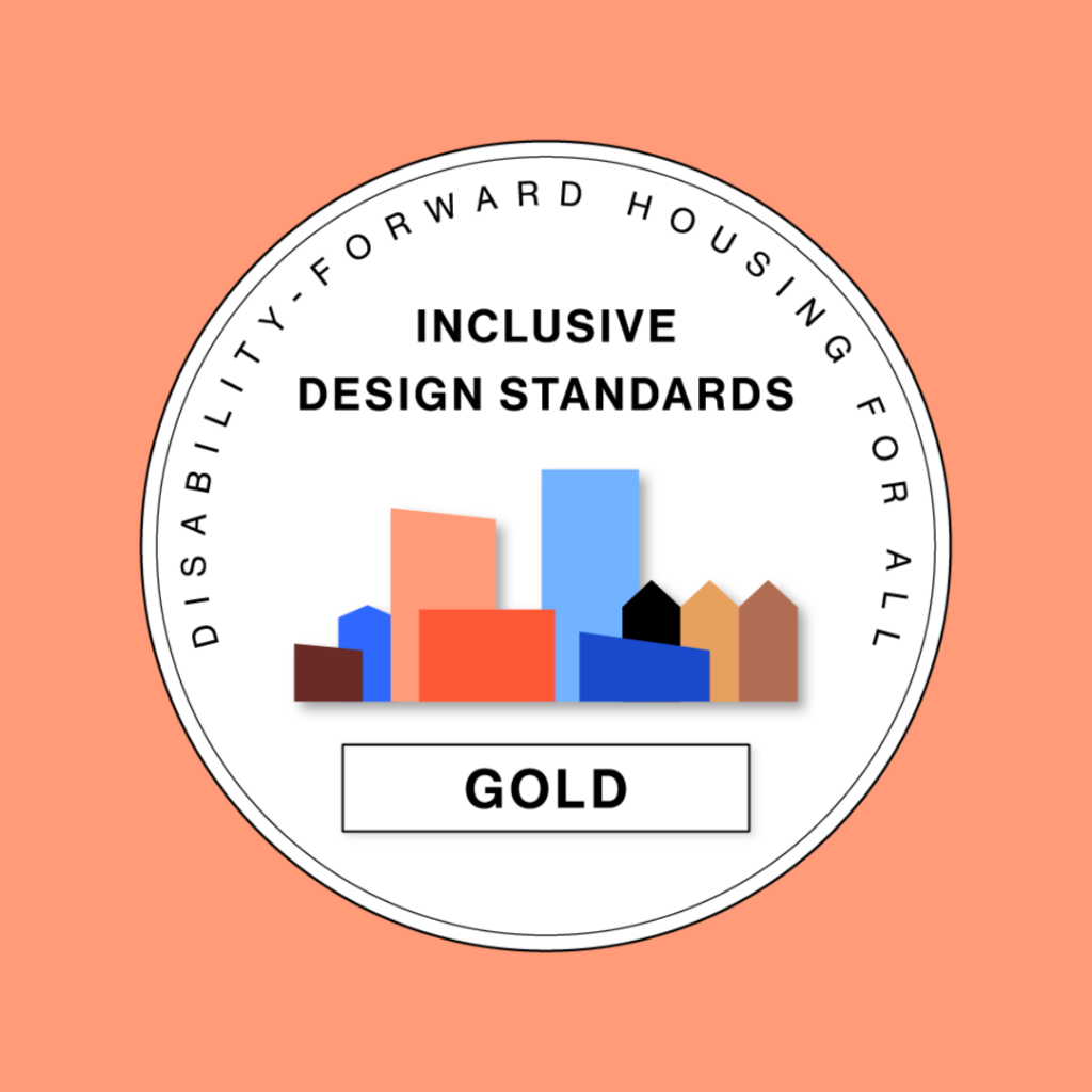 A circular emblem with the words “Disability-forward housing for all” around the outer ring. In the center, bold text reads “Inclusive design standards.” Below the text is a row of colorful geometric building shapes in shades of red, orange, blue, and brown, representing a community skyline. Beneath the buildings is a rectangular box with the word “Gold” in bold black letters, signifying the project’s certification level. The background is coral.
