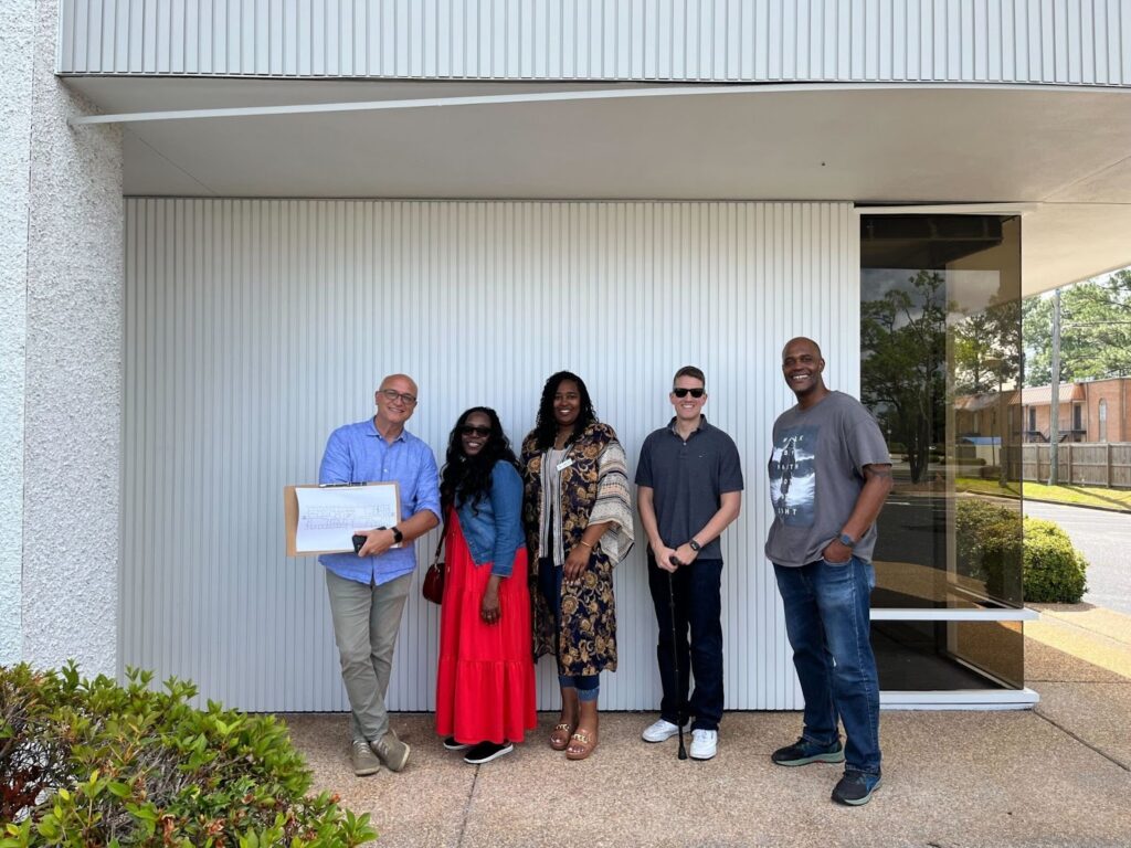 Five people of different races, genders, and with and without disabilities, stand and smile outside a white building. One person holds a large paper and a clipboard with building schematics on it.