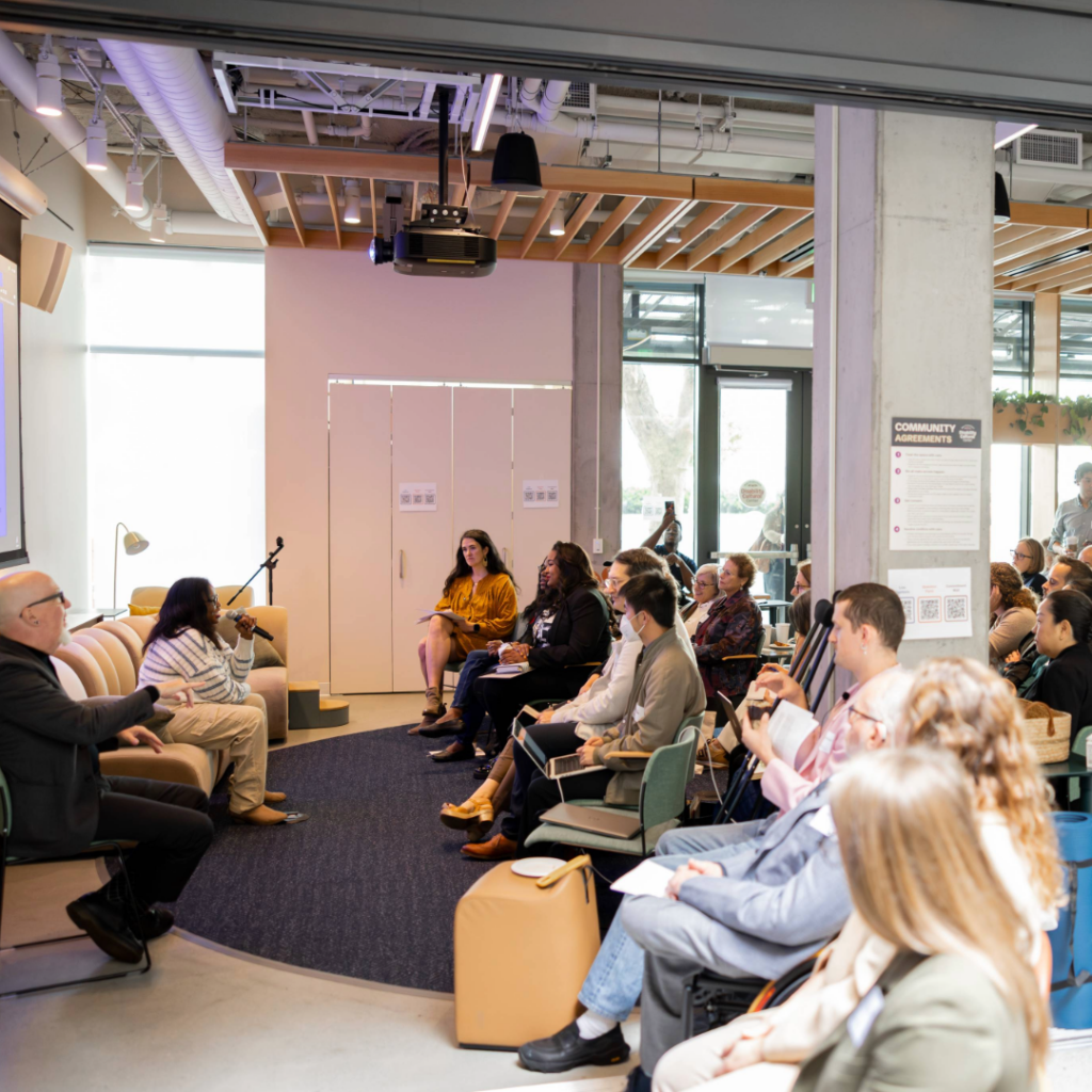 A session during the Disability-Forward Housing Futures Summit. A diverse group of adults sits in a semicircle inside a modern, well-lit community space. Keah Brown, a Black person with long straight hair, wearing a white and gray striped sweater and khaki pants, sits at the front of the room and speaks into a microphone. A sign language interpreter also sits at the front of the room. Attendees listen attentively, some taking notes, with a projector mounted overhead and large windows and exposed wood beams visible in the background.