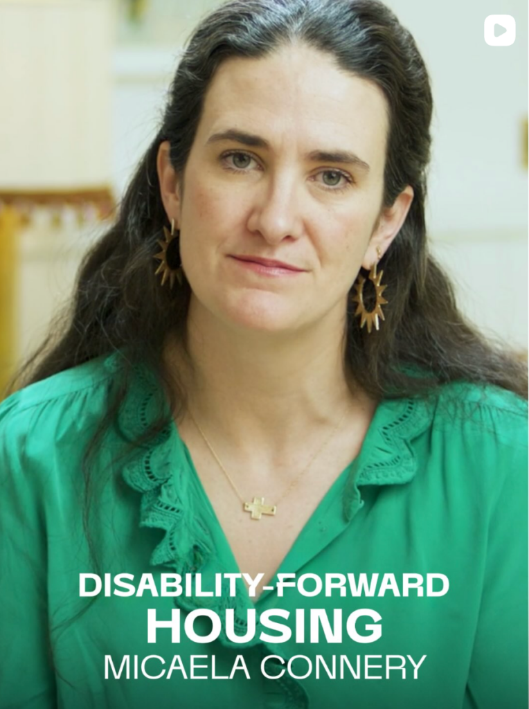 A still image from Micaela Connery’s Brief But Spectacular segment. Micaela looks at the camera with a slight smile. She is a white woman with long, brown hair, green eyes, wearing a green blouse and gold jewelry, including a cross necklace and sun-shaped earrings. The text “Disability-Forward Housing: Micaela Connery,” is in the lower third of the image.
