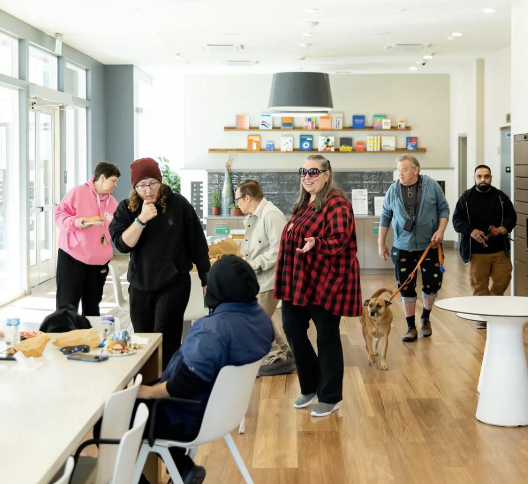 A group of The Kelsey Ayer Station residents gathers and talks in the lobby. Some sit while others stand and walk through the lobby. The lobby is a bright, modern indoor common area with light wood floors and white walls. The mailroom is visible in the background with books displayed on shelves.