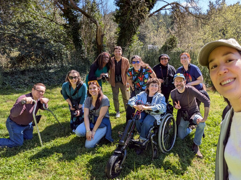Members of our team, people with and without disabilities of different races and genders, take a selfie with two bird-watching guides. The team poses in a group on the grass, with trees visible behind them.