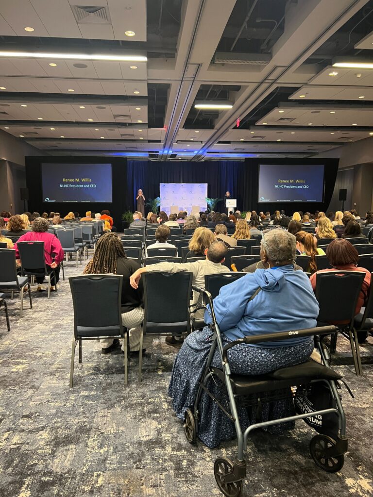 A conference room of seated participants watching a session at the NLIHC Policy Forum. Two screens and two speakers stand on stage at the front of the room.