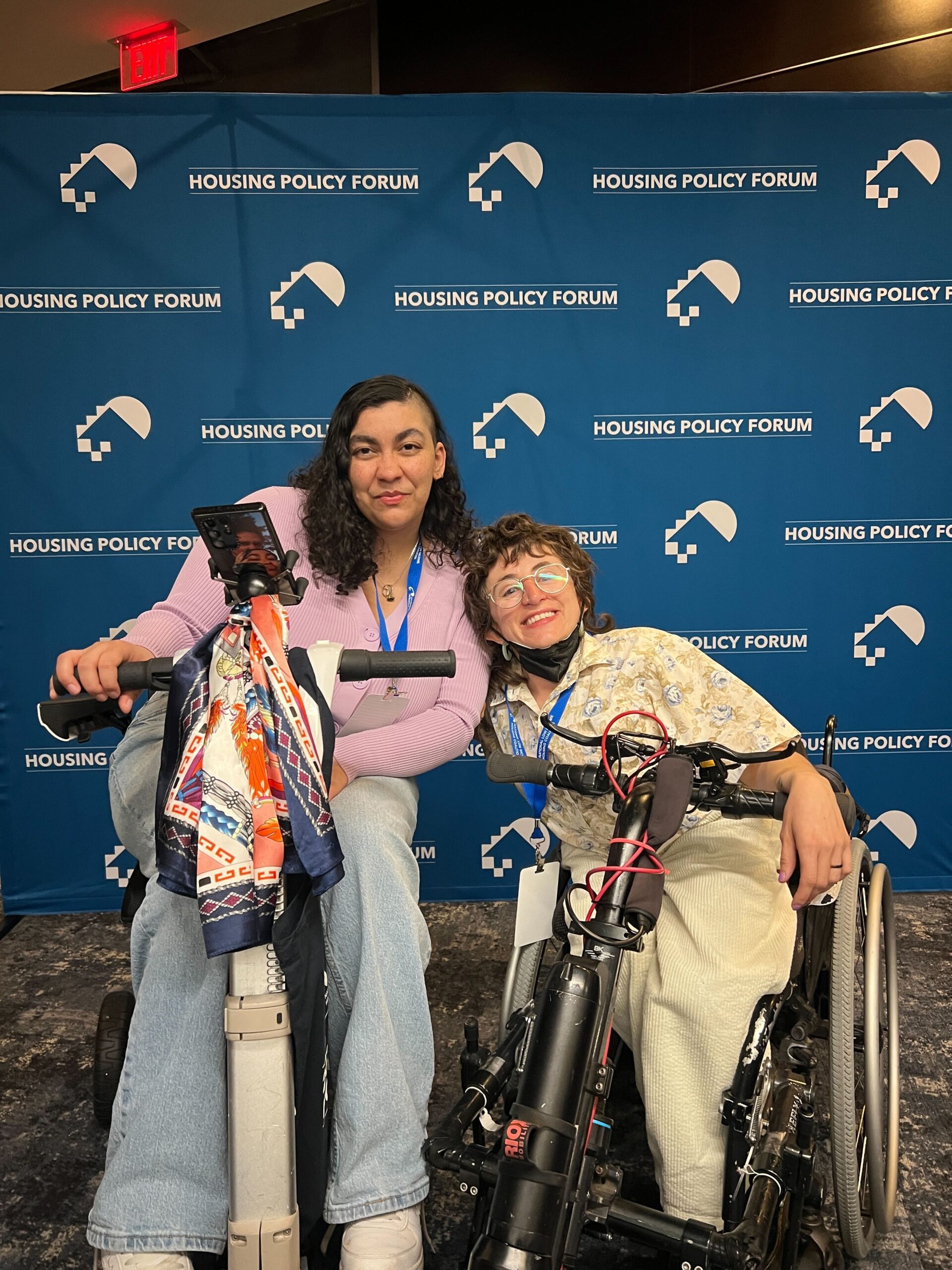 Allie is pictured with Iliana, the Housing Organizer at Access Living and a former Raise the Roof participant. Both of them are in power wheelchairs. The words “Housing Policy Forum” and the logo of The National Low Income Housing Coalition are pictured on a backdrop behind them. They are both wearing lanyards and casual clothing.