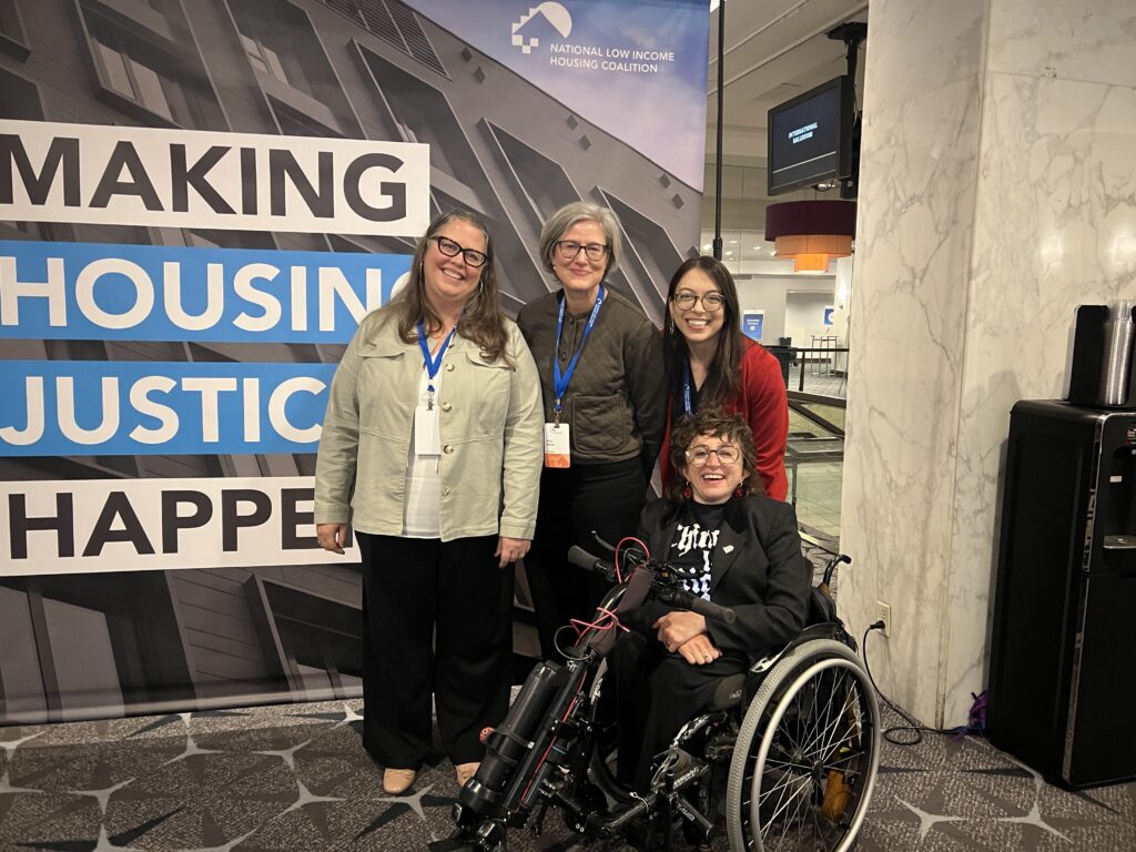 Allie, our Managing Director, poses with three partners in front of a backdrop reading “Make Housing Justice Happen” and featuring the National Low Income Housing Coalition logo. Allie is pictured in a power wheelchair, with the three partners standing behind her. They all wear casual clothing and lanyards and are smiling at the camera.