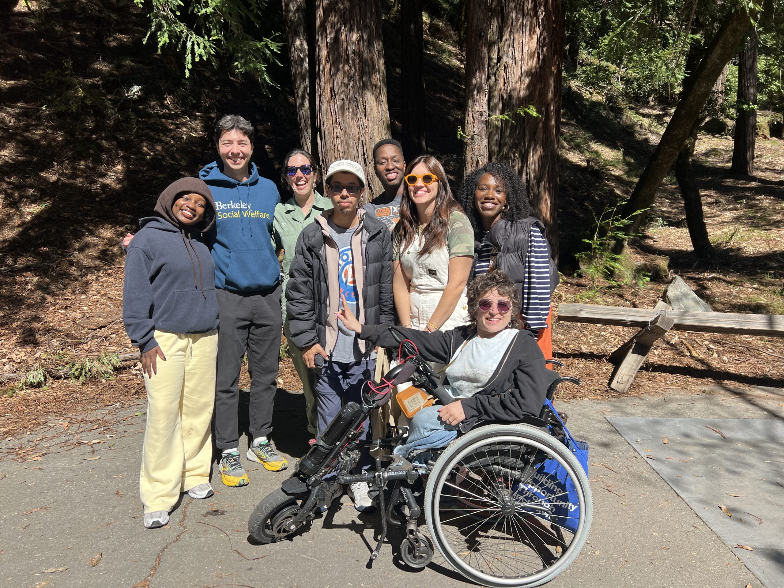 Members of our team, people with and without disabilities of different races and genders, take a selfie with two bird-watching guides. The team poses in a group on the grass, with trees visible behind them.