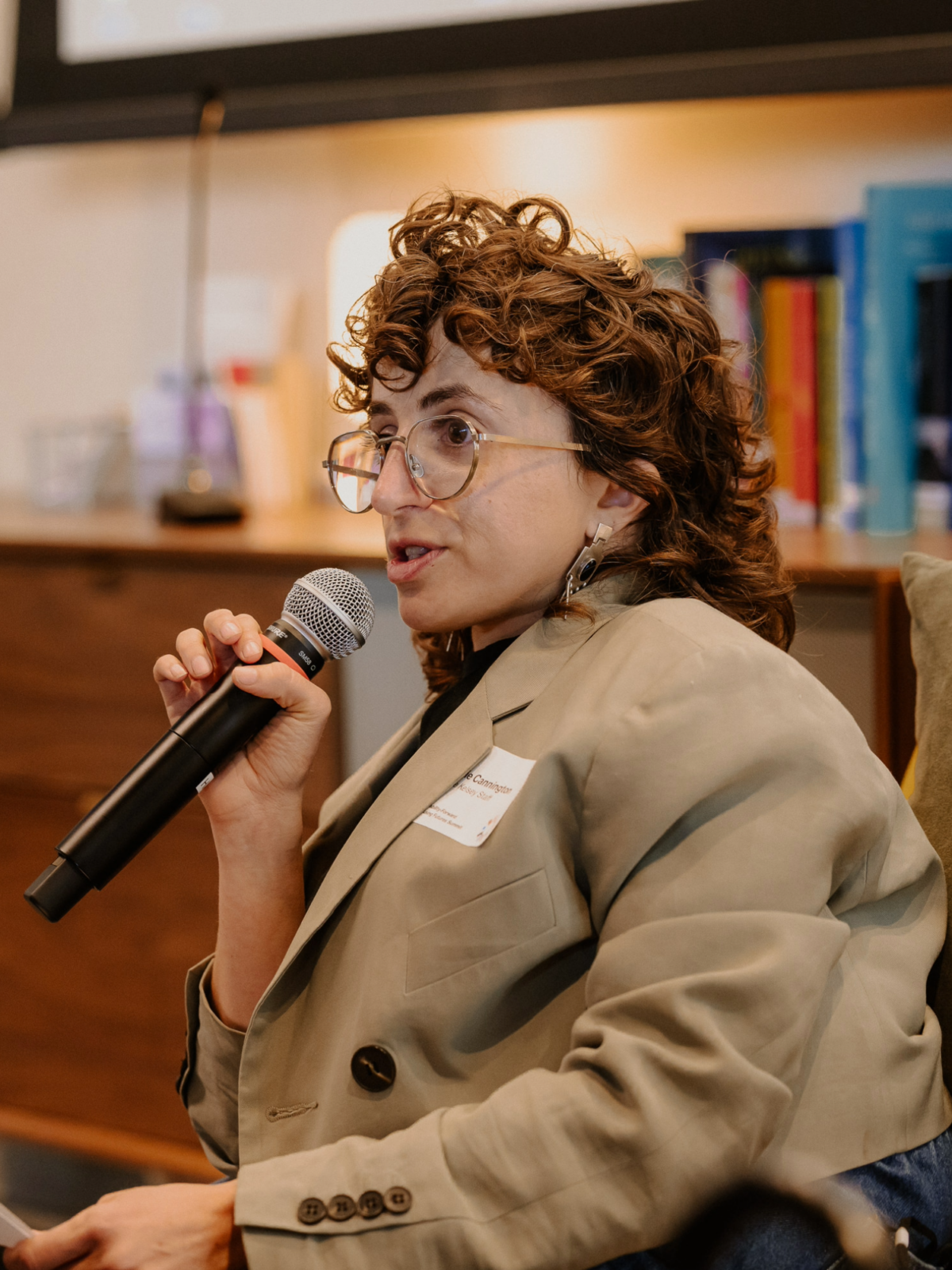 A close-up photo of Allie, a white queer person with short curly hair and glasses, holding a microphone and speaking. They are wearing a beige blazer and silver dangling earrings.