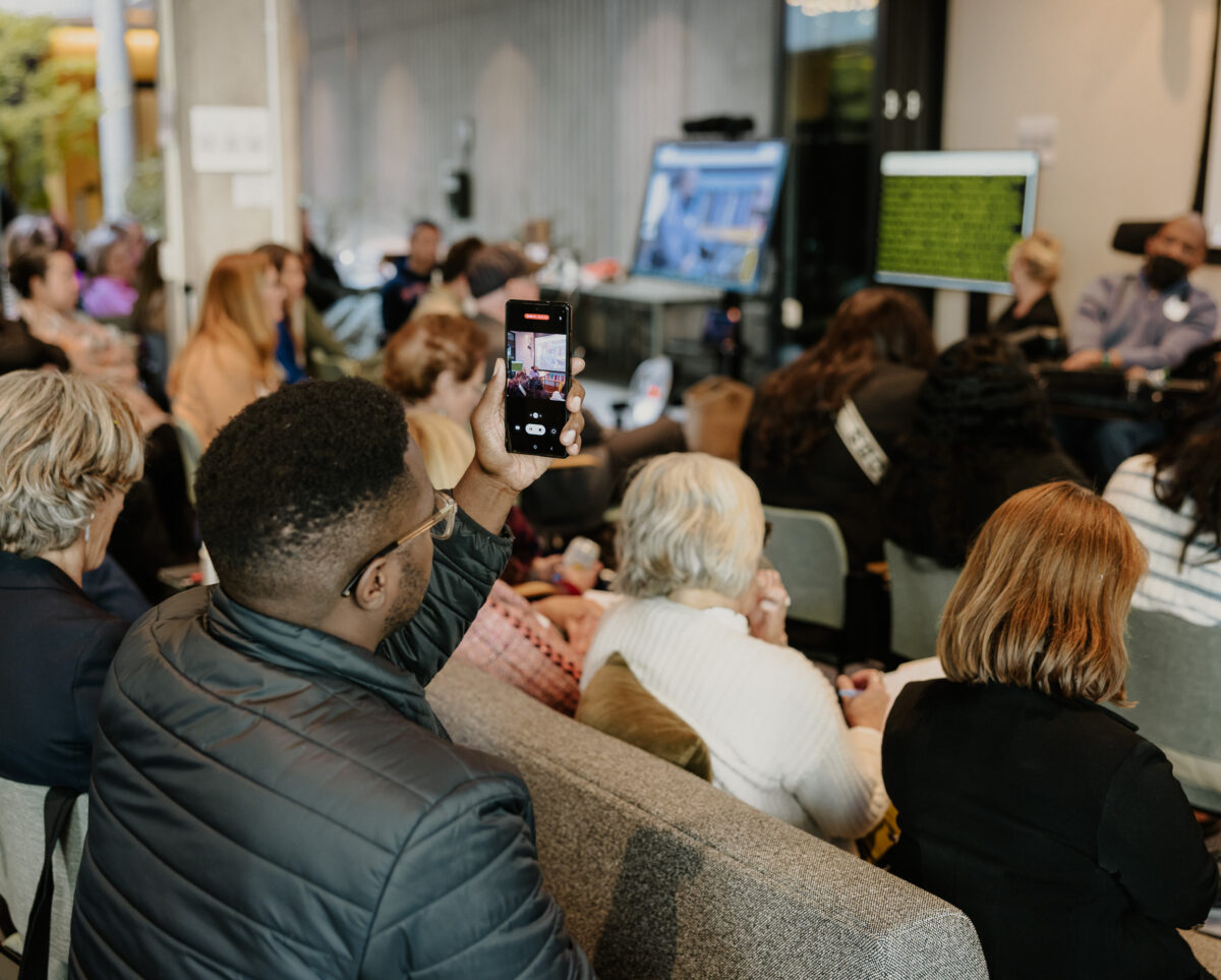 People watch a presentation at the Disability-Forward Housing Futures Summit. Out of focus, a man in a wheelchair presents at the front of the room. In focus, an audience member records the presentation on their phone.