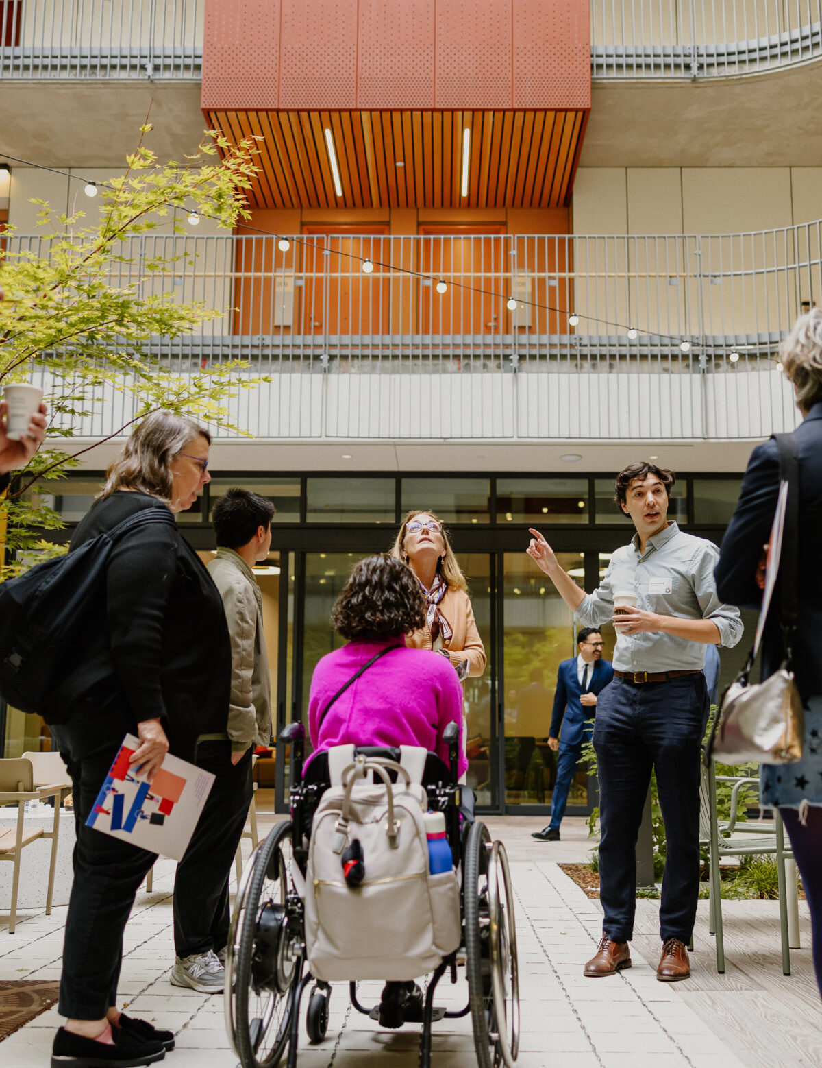 A group of people with and without disabilities on a tour in The Kelsey Civic Center’s courtyard. They are gathered in a loose circle, with one person gesturing and talking. One tour member gazes up at the balconies surrounding the courtyard, and other tour members hold coffee cups and The Kelsey folders.