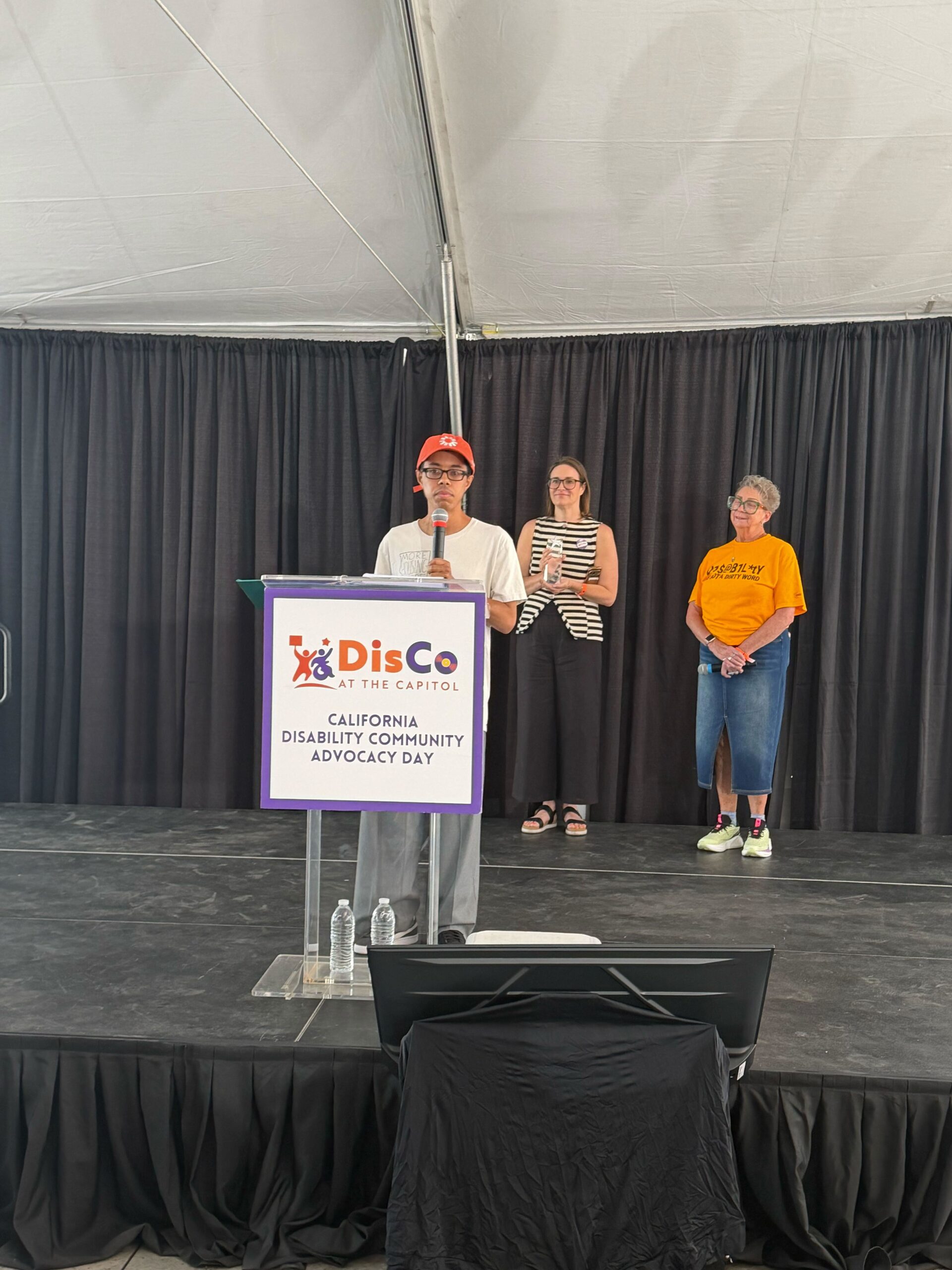 Isaac, a Black man wearing a white t-shirt and an orange The Kelsey baseball cap, stands behind a podium on a stage and speaks into a microphone. Two people stand behind him. One is holding a glass award. A sign on the podium reads “DisCo at the Capitol: California Disability Community Advocacy Day.”