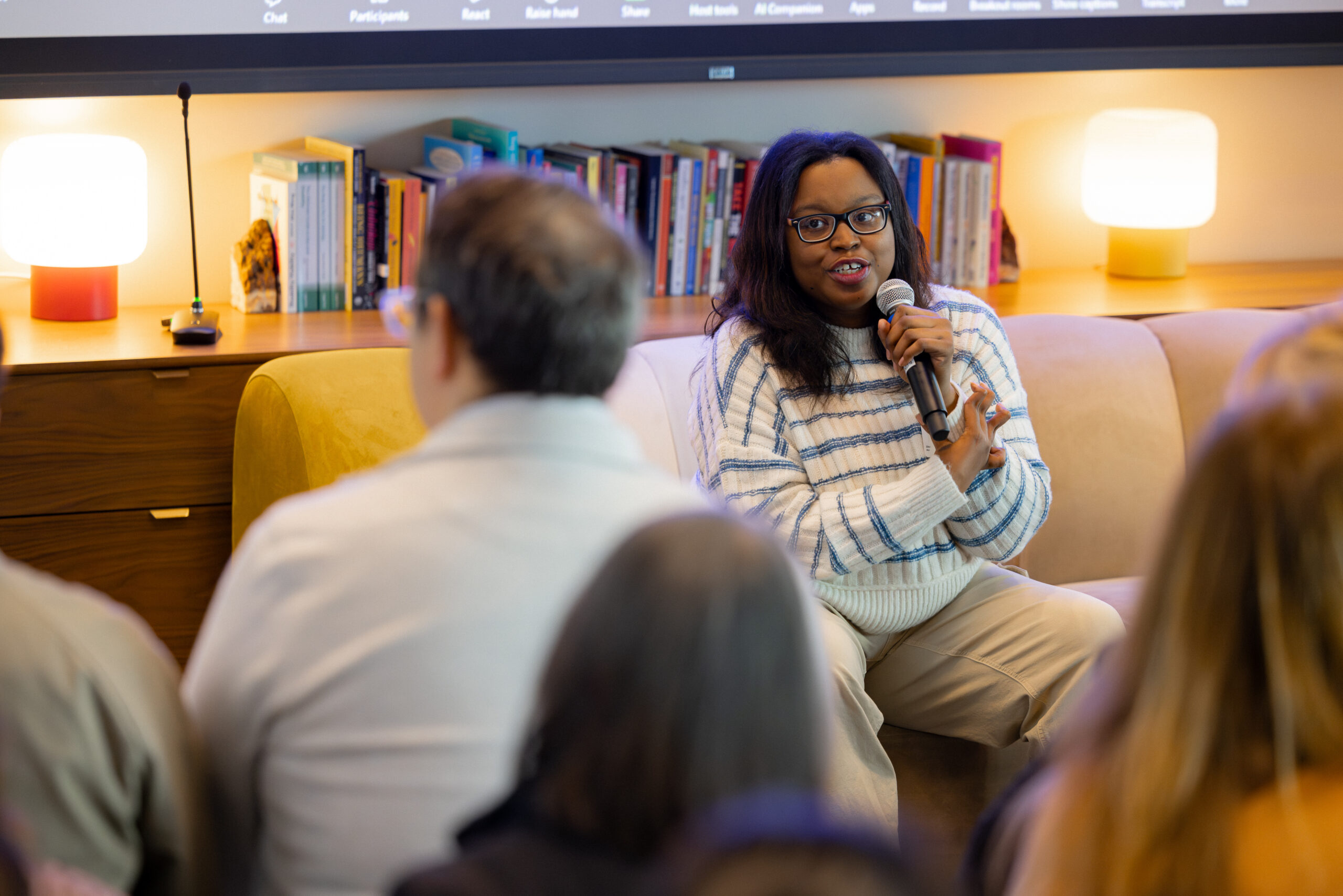 Keah Brown, a Black woman with long, dark brown hair and glasses, wearing a white-and-blue striped shirt, holds a microphone and speaks to an audience. The audience is out of focus, and the camera focuses on her.