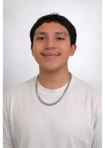 Image Description: A young person with short dark hair smiles at the camera, wearing a white shirt, silver chain, small earrings, and an eyebrow piercing against a softly plain background.