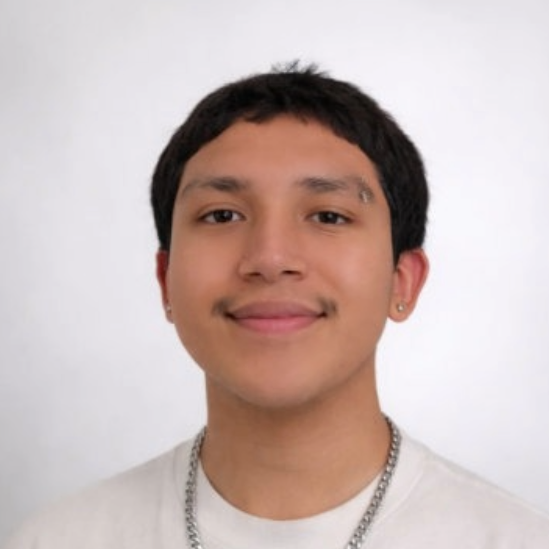 A young person with short dark hair smiles at the camera, wearing a white shirt, silver chain, small earrings, and an eyebrow piercing against a softly plain background.