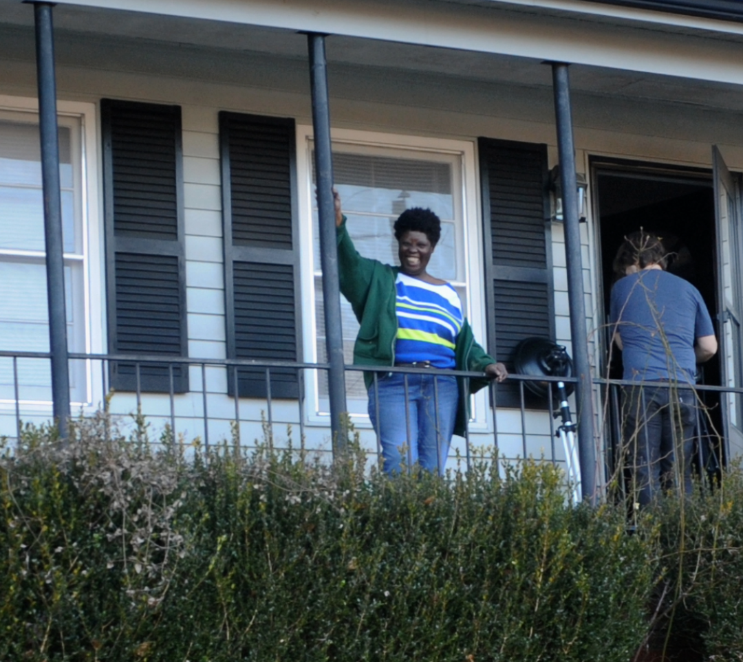 Lois Curtis, a Black woman with short, curly hair, wearing a green sweatshirt, a blue striped shirt, and jeans, stands on the front porch of a house. She is smiling widely, with one hand on the porch railing and the other on a beam. A person behind her opens the house door, their back to the camera.