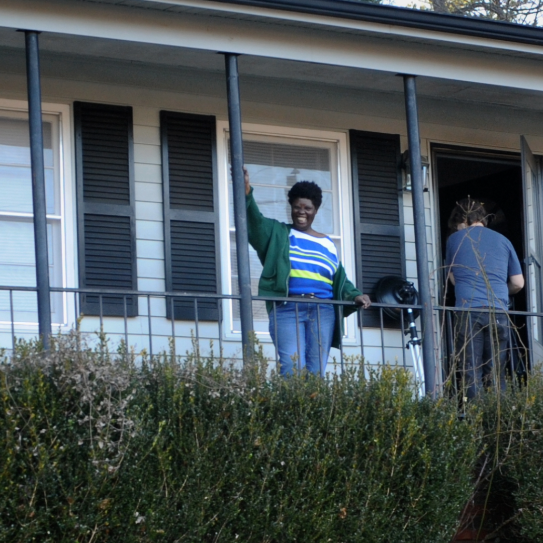 Lois Curtis, a Black woman with short, curly hair, wearing a green sweatshirt, a blue striped shirt, and jeans, stands on the front porch of a house. She is smiling widely, with one hand on the porch railing and the other on a beam. A person behind her opens the house door, their back to the camera.