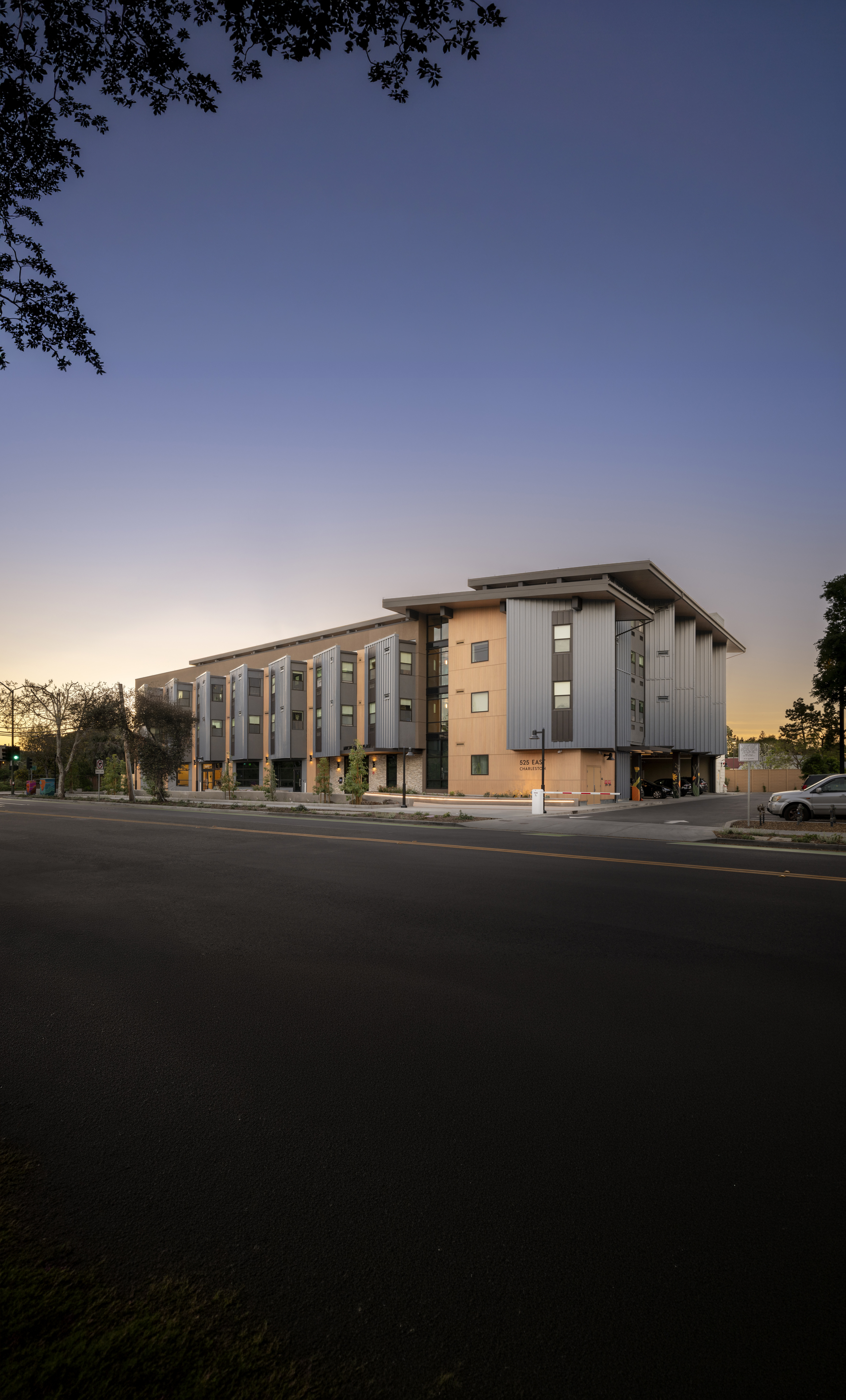 A picture of a modern multi-story apartment building at dusk, viewed from across a dark, empty street. The building features a contemporary architectural style with gray metal and warm wood-toned panels, and a flat, overhanging roofline. The sky above transitions from a deep twilight blue at the top to a warm orange glow near the horizon behind the building. Tree branches frame the very top of the image, and a few parked cars are visible on the far right.