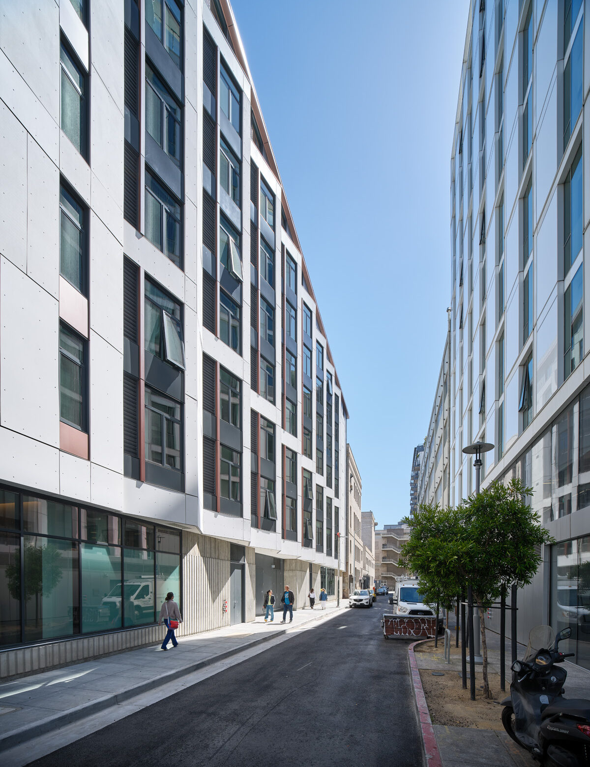 A bright, quiet city street lined by two modern apartment buildings. The buildings rise five to six stories on either side, forming a canyon-like corridor. A few people walk along the sidewalks, young trees dot the street edge, and a scooter and several vehicles are parked nearby.