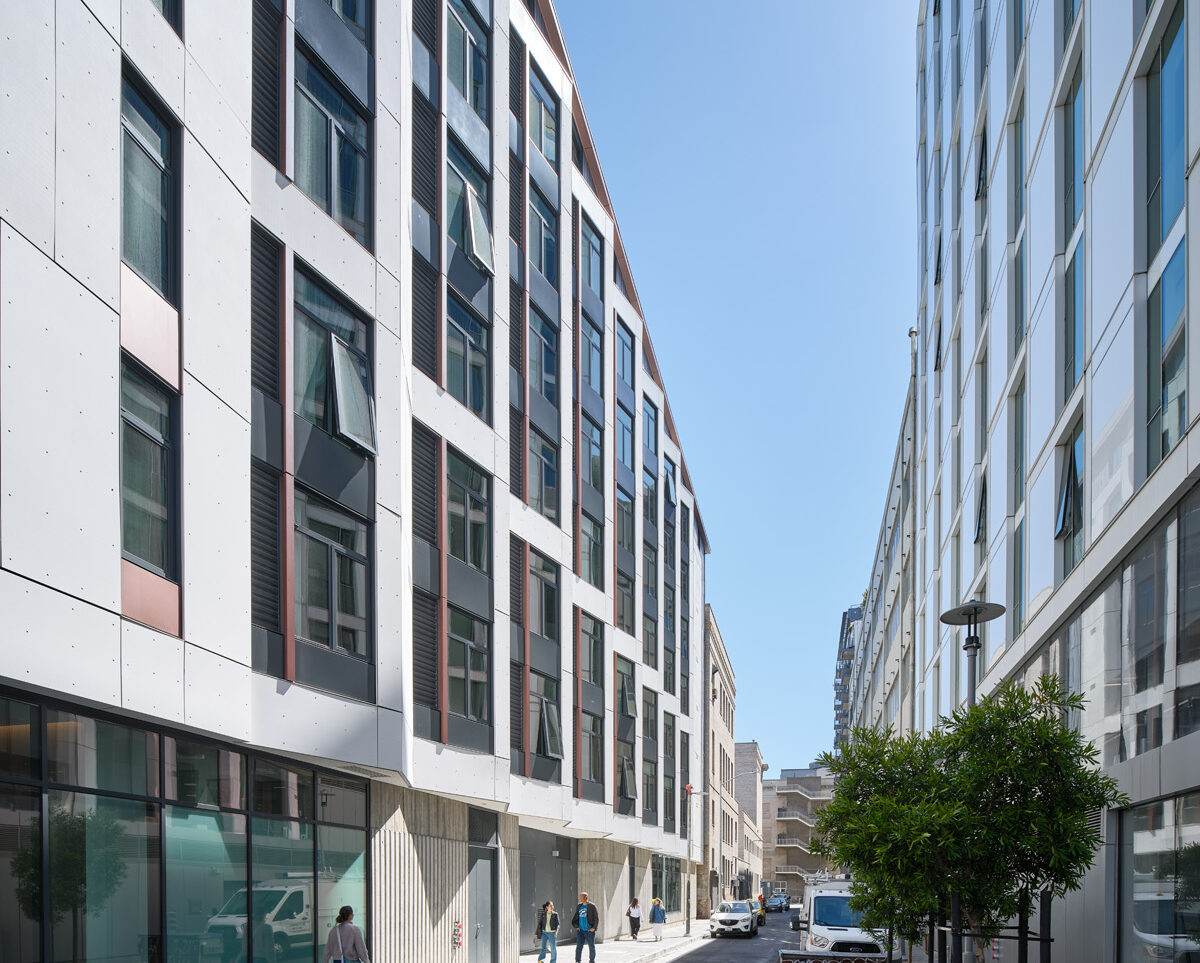 A bright, quiet city street lined by two modern apartment buildings. The buildings rise five to six stories on either side, forming a canyon-like corridor. A few people walk along the sidewalks, young trees dot the street edge, and a scooter and several vehicles are parked nearby.