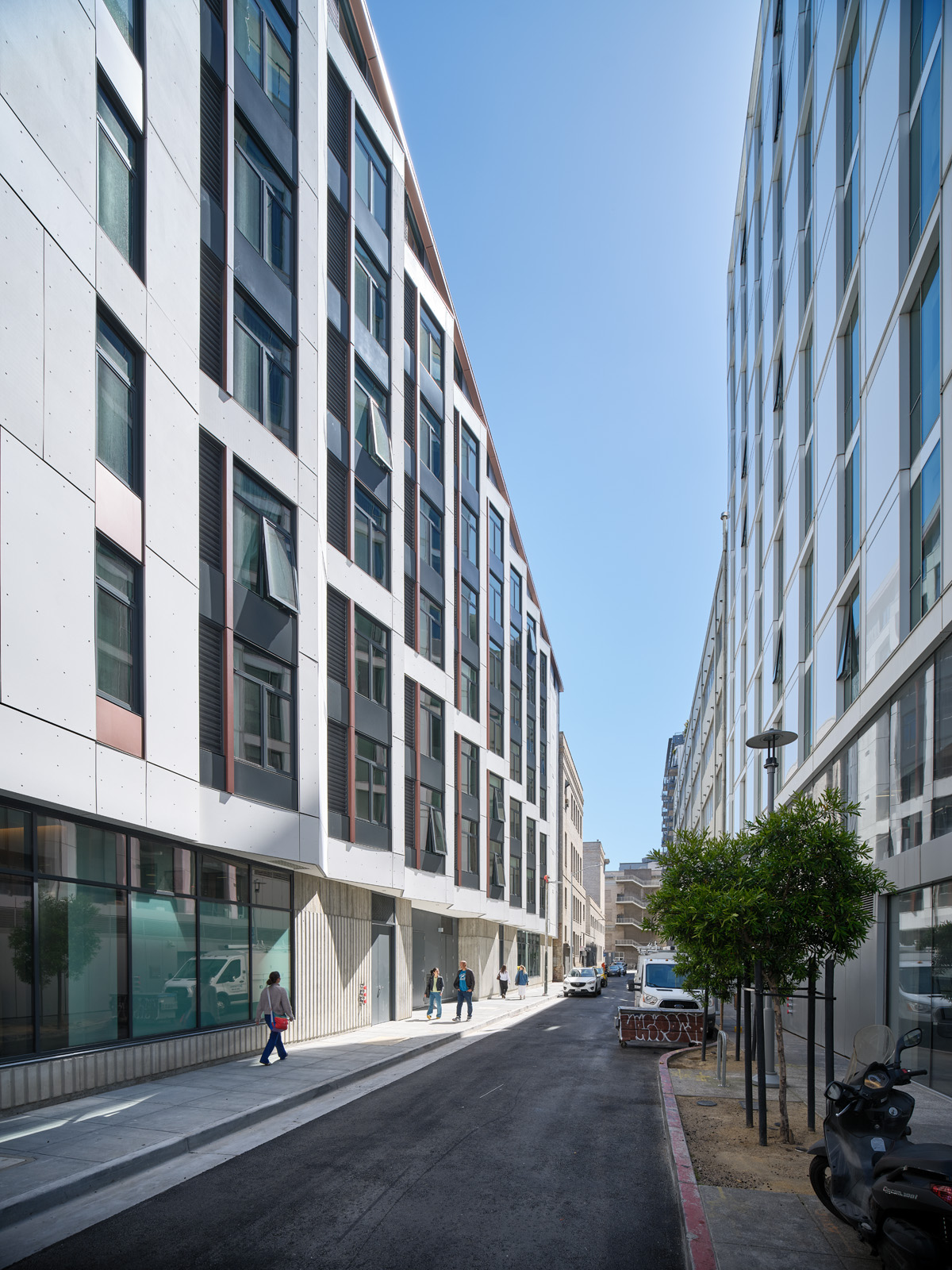 A bright, quiet city street lined by two modern apartment buildings. The buildings rise five to six stories on either side, forming a canyon-like corridor. A few people walk along the sidewalks, young trees dot the street edge, and a scooter and several vehicles are parked nearby.
