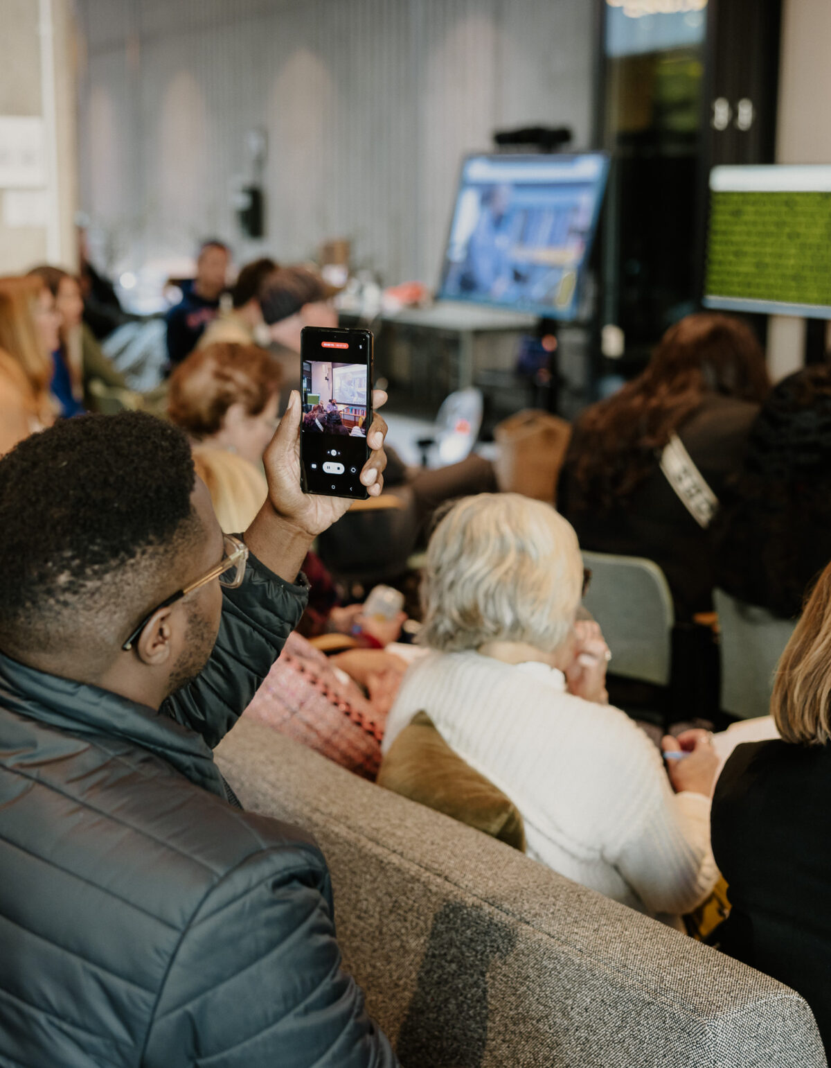 People watch a presentation at the Disability-Forward Housing Futures Summit. Out of focus, a man in a wheelchair presents at the front of the room. In focus, an audience member records the presentation on their phone.