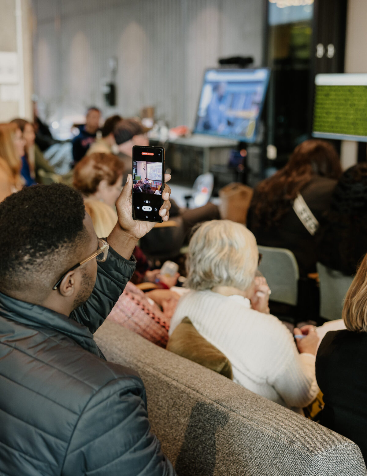 People watch a presentation at the Disability-Forward Housing Futures Summit. Out of focus, a man in a wheelchair presents at the front of the room. In focus, an audience member records the presentation on their phone.