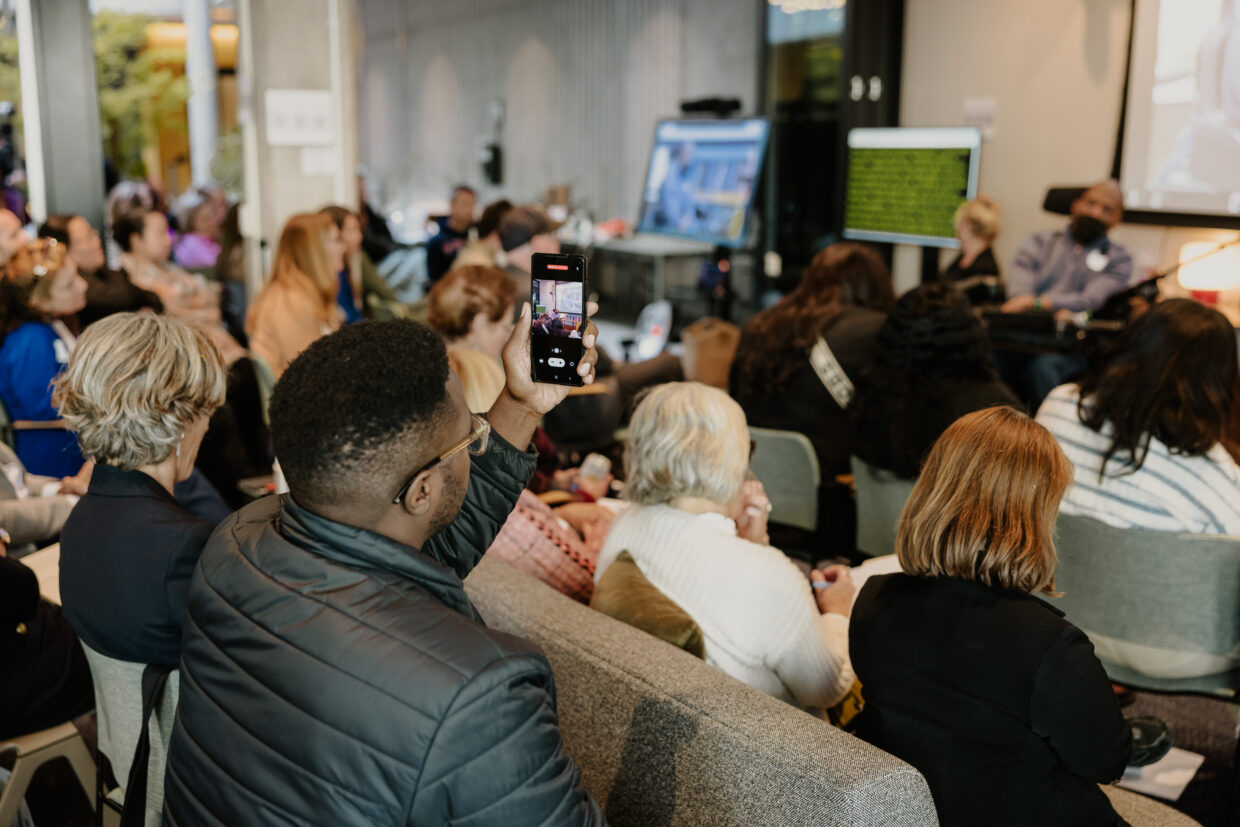 People watch a presentation at the Disability-Forward Housing Futures Summit. Out of focus, a man in a wheelchair presents at the front of the room. In focus, an audience member records the presentation on their phone.