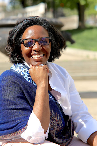 Angela Gravely Smith, a woman smiling with black-brown hair with streaks of gray. She is wearing navy blue glasses, thin-hooped silver earrings, and a white shirt with a blue scarf. She is sitting down outside, resting her chin on her hand. The background is blurred.