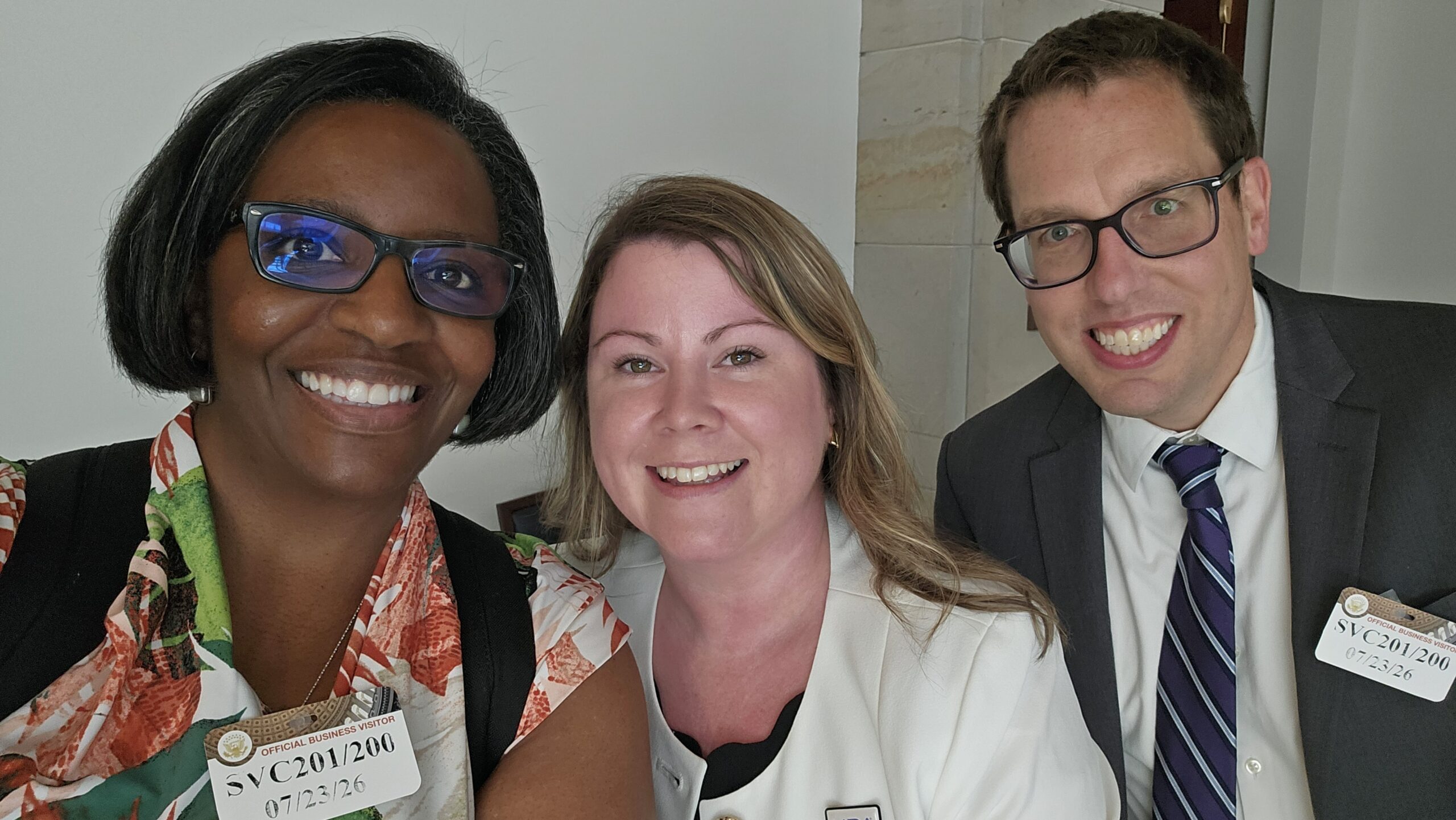 Angela, our Managing Director of Technical Assistance, takes a selfie with two people at the CCD Briefing on Community Living. Angela, far left, is a woman with a short dark bob haircut, glasses, smiling, and wearing an orange, white, and green patterned blouse with a visitor badge clipped on it. To her right, a person with long blonde hair and light eyes smiles, wearing a white blazer. At the far right, a smiling person with short dark brown hair and glasses, wearing a coat and tie with a visitor badge, leans into the picture.