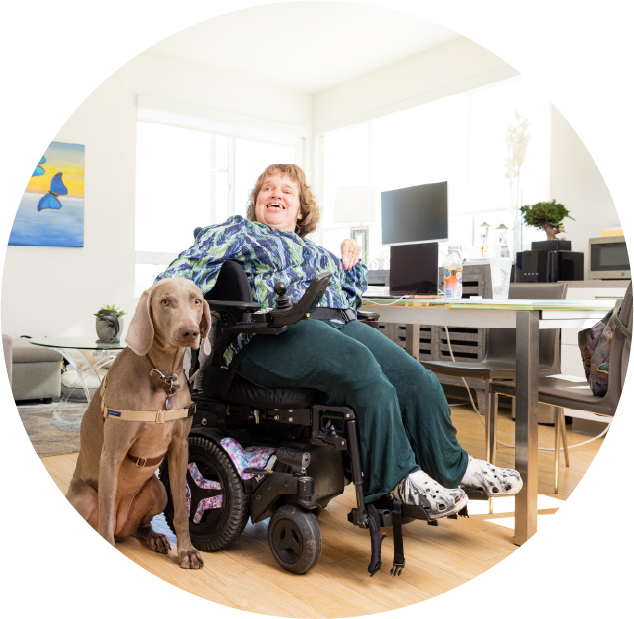 Woman in a wheelchair next to her dog and sitting by a table with a computer on top of the table