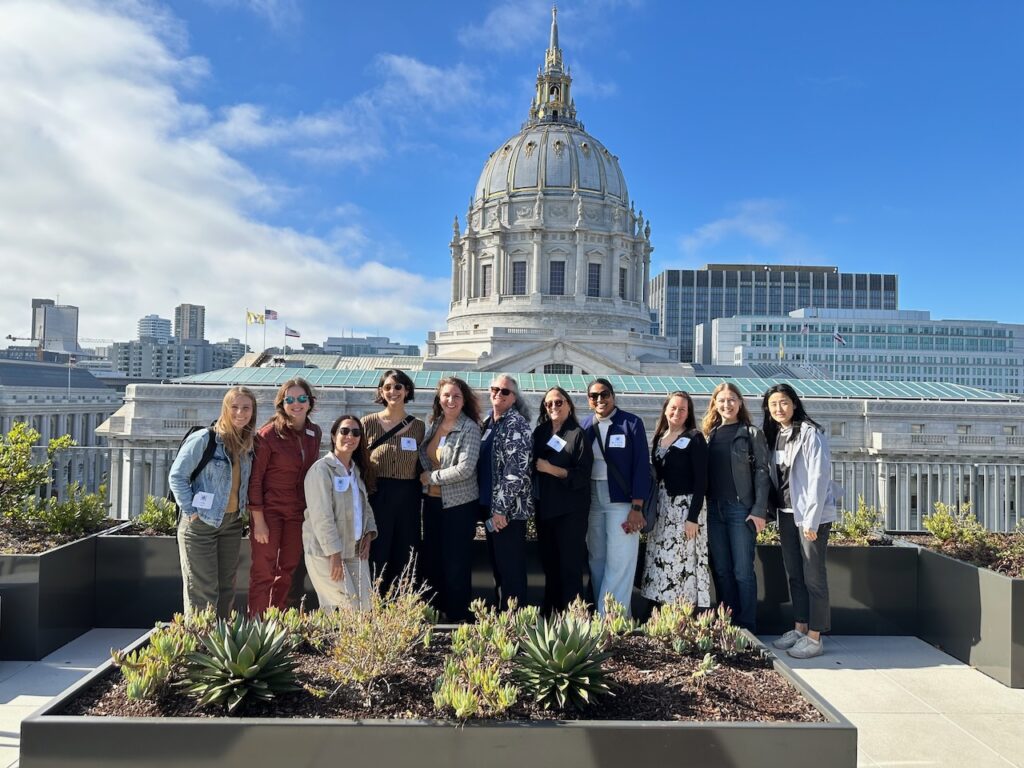 Members of the Women’s Development Collaborative pose on the roof of The Kelsey Civic Center with the dome of San Francisco City Hall in clear view behind them. They wear casual clothing, name tags, and some have sunglasses on. On the rooftop, they are surrounded by garden beds with various small green plants.